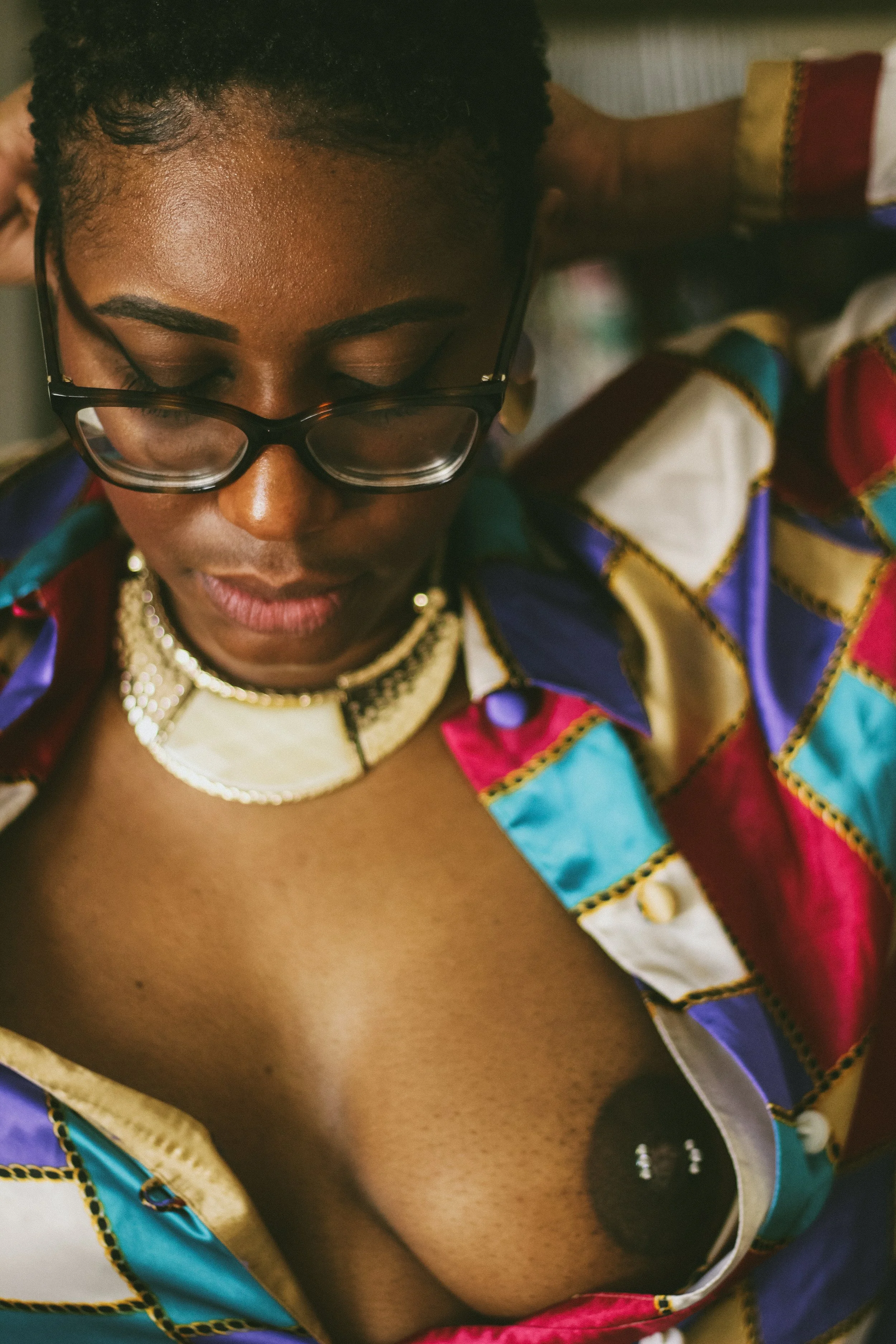 A woman with short curly hair wearing glasses and colorful, patterned clothing, looks down with her eyes closed.