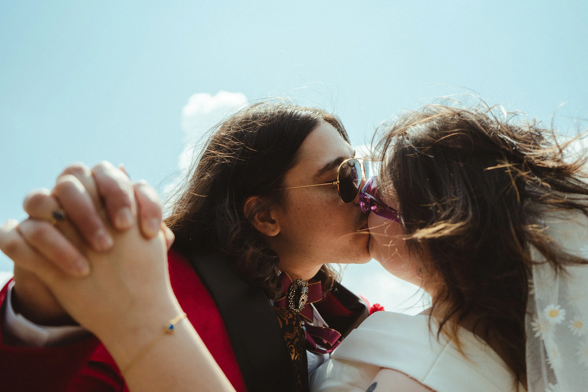 Two women embrace in a close hug against a blue sky with a few clouds. One woman has dark hair and wears sunglasses, while the other has curly hair. The women are holding hands and appear to be sharing an emotional moment.