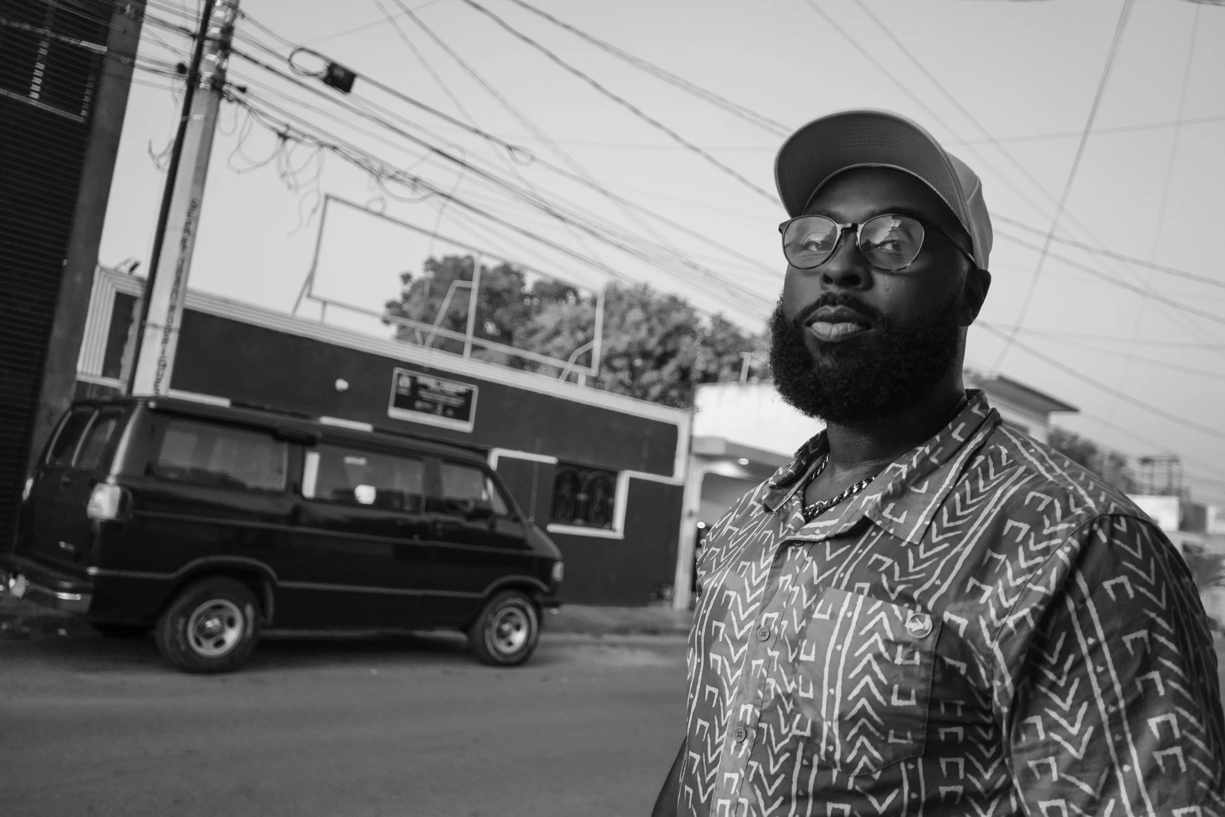 A black and white photo of a man with glasses and a cap, standing outdoors in front of a black vehicle, with electrical wires and trees visible in the background.