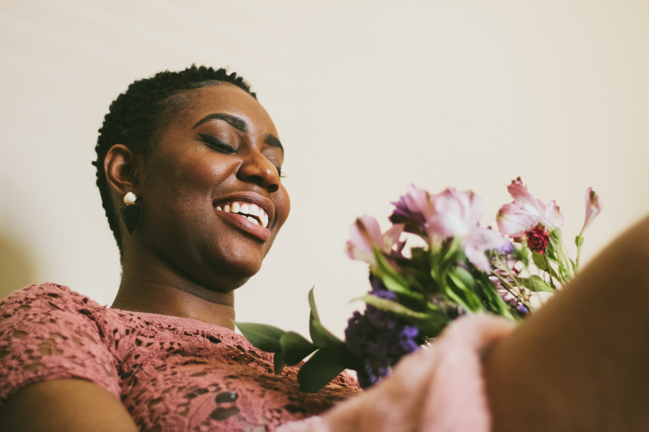 A woman with short dark hair, earrings, and wearing a pink lace top, smiling and holding a bouquet of pink and purple flowers.