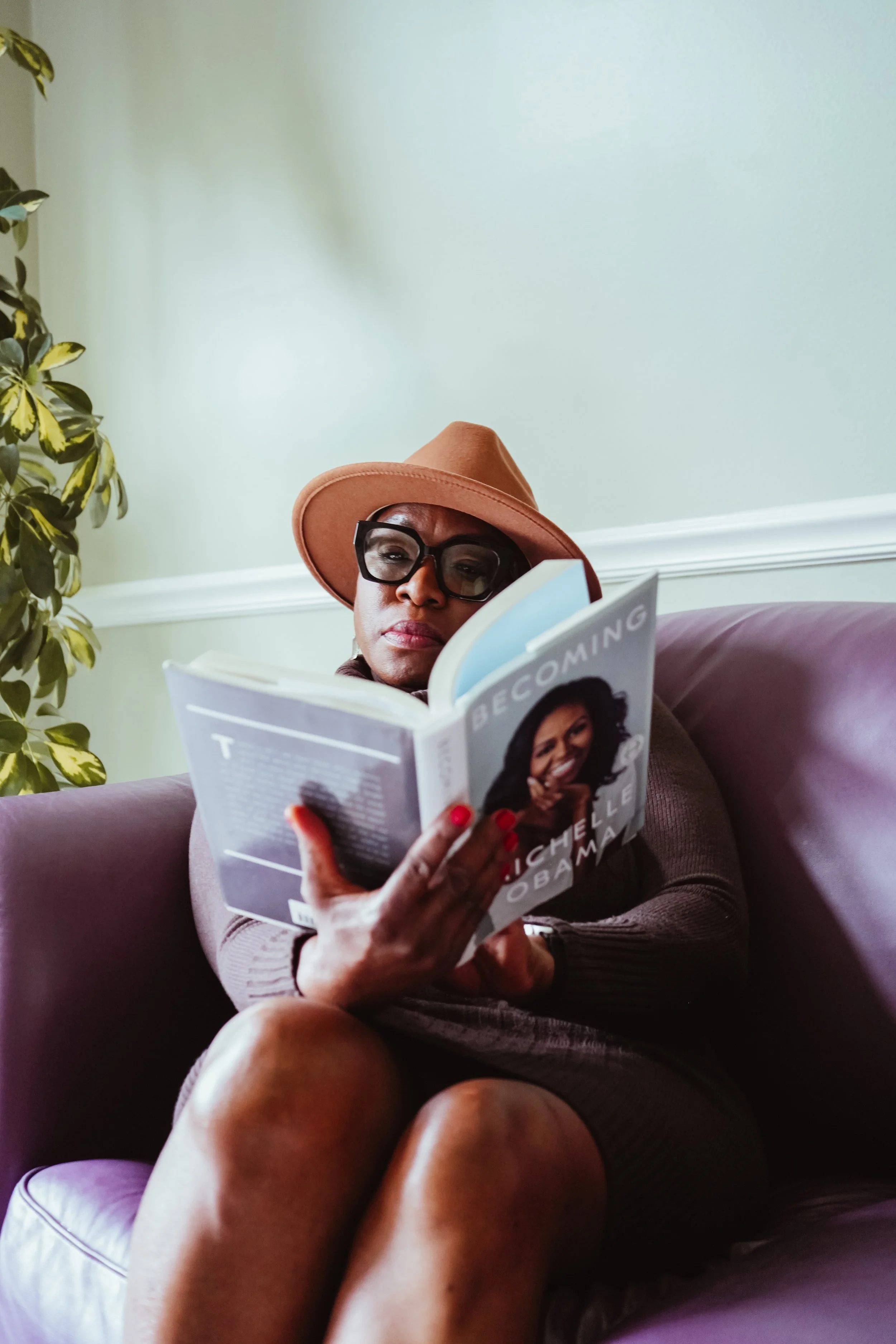 A woman wearing a brown hat and black glasses sitting on a purple couch, reading a book titled "Becoming" by Michelle Obama, with a green wall and a potted plant in the background.