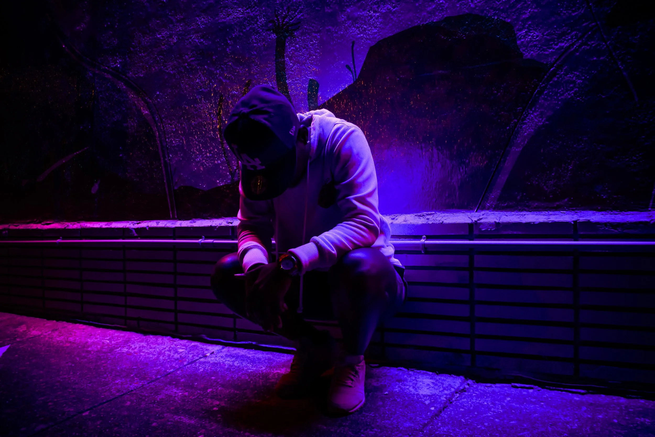 A person sitting on a low wall in a dark environment with vibrant purple and pink lighting. They are wearing a hoodie, cap, and sneakers, with their head bowed and face hidden, under a glowing purple and blue ceiling.
