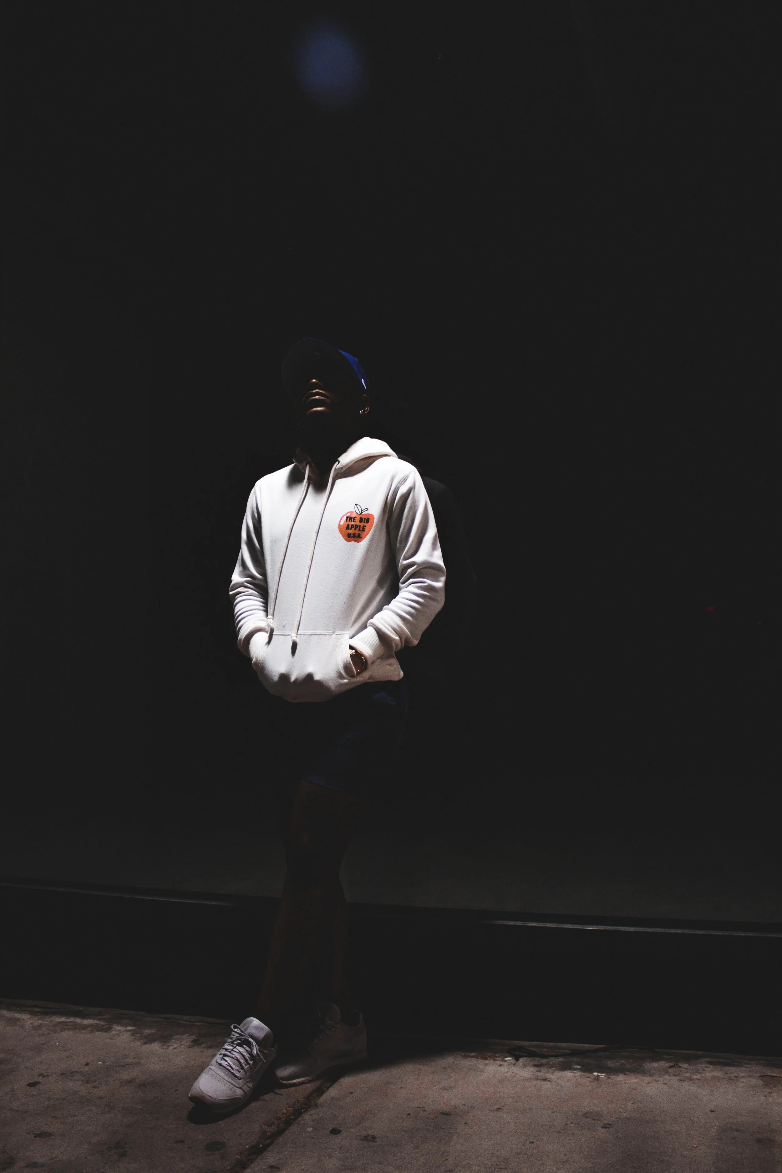 A person stands in a dark space, wearing a white hoodie with a graphic that says 'The Big Apple USA', black shorts, and white sneakers. The person's face is partially shadowed, and they look upwards.