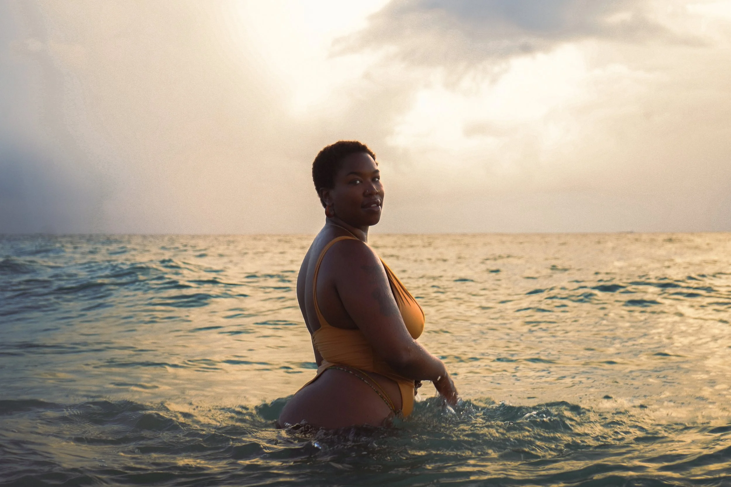 A woman with short hair and earrings standing in the ocean at sunset, looking over her shoulder.