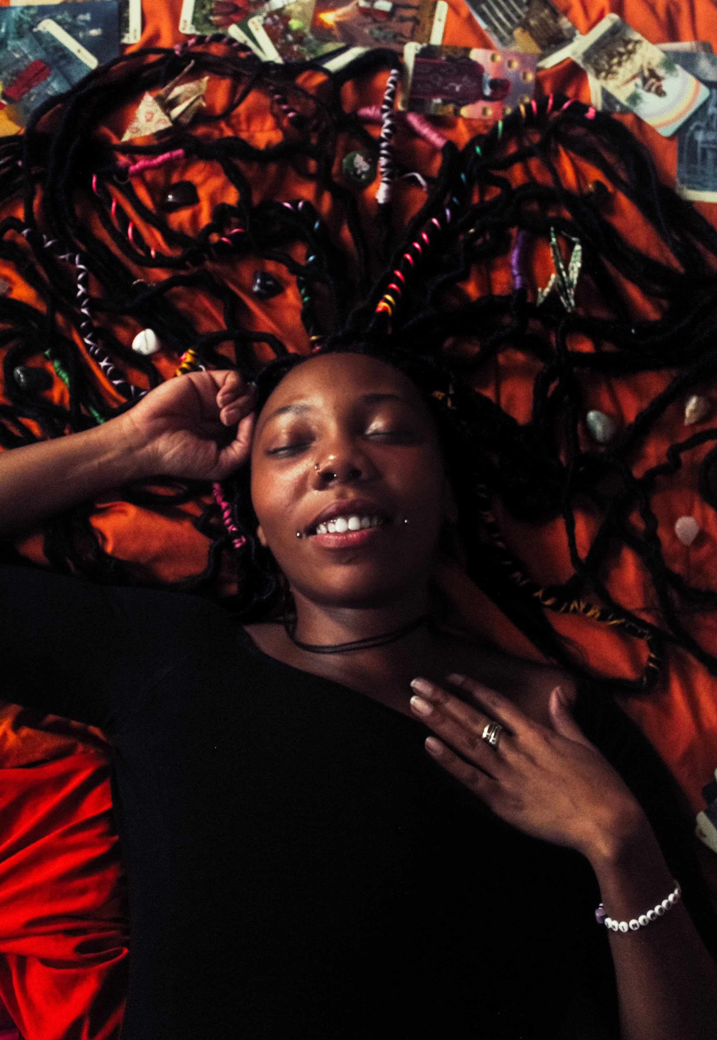 A person with dark braided hair with colorful beads, lying on an orange fabric with tarot cards scattered around. They have dark skin, closed eyes, and a nose piercing, wearing a black shirt, a beaded bracelet, and rings on their fingers.