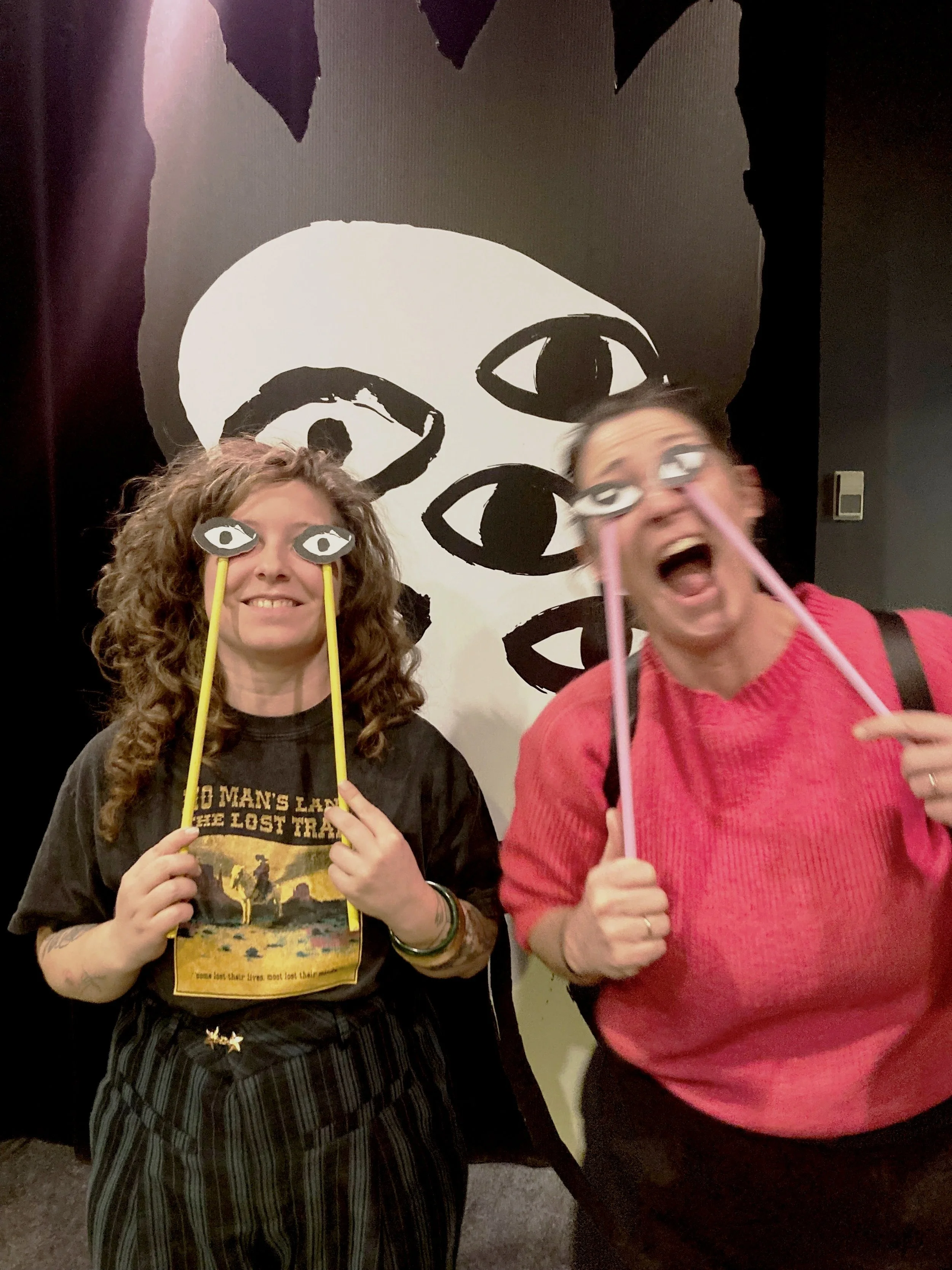 Deux femmes posant devant un mur noir avec une grande illustration d'un visage en noir et blanc, portant des accessoires avec des yeux.»