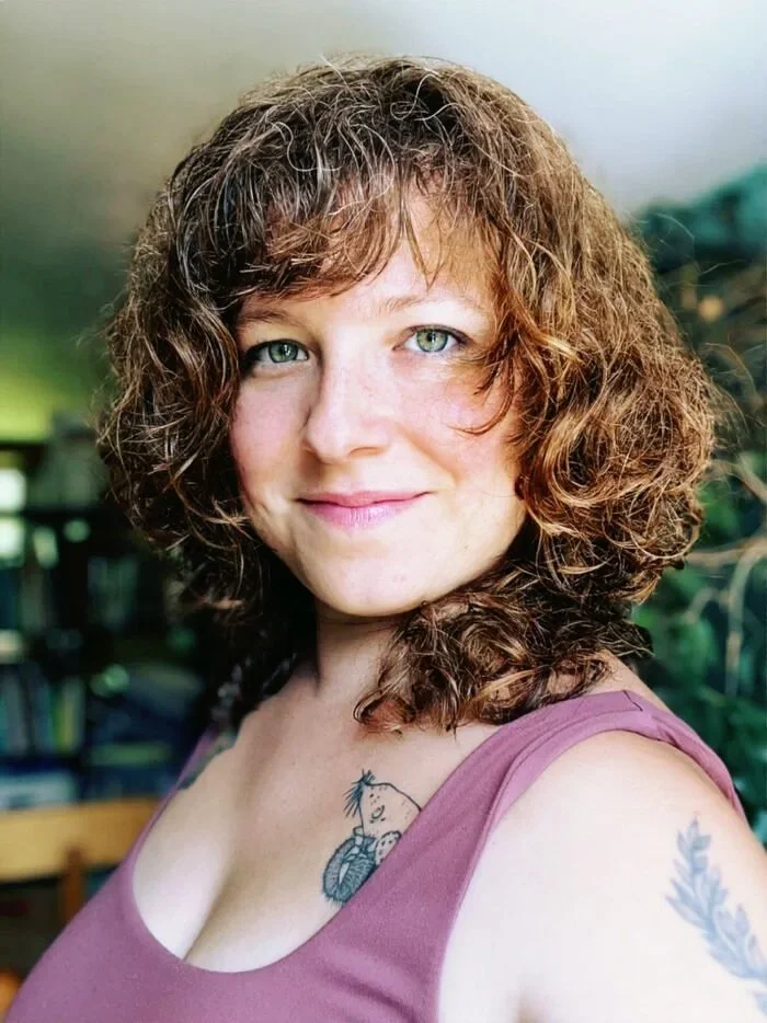 Une femme avec des cheveux bouclés et bruns, portant un haut mauve, affichant un sourire, avec des tatouages visibles sur l'épaule et la poitrine, dans un environnement naturel ou intérieur avec des plantes.