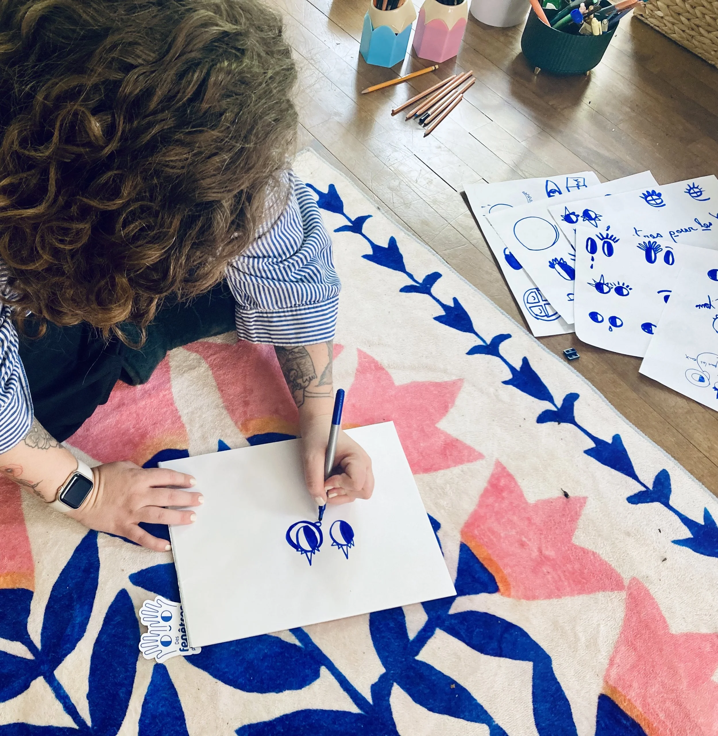 Une personne avec des cheveux bouclés, portant une tenue à rayures, assise sur un tapis, en train d'écrire ou de dessiner sur une feuille blanche. Sur la table en bois devant elle, il y a plusieurs feuilles avec des dessins bleus, un stylo et des crayons de couleur.