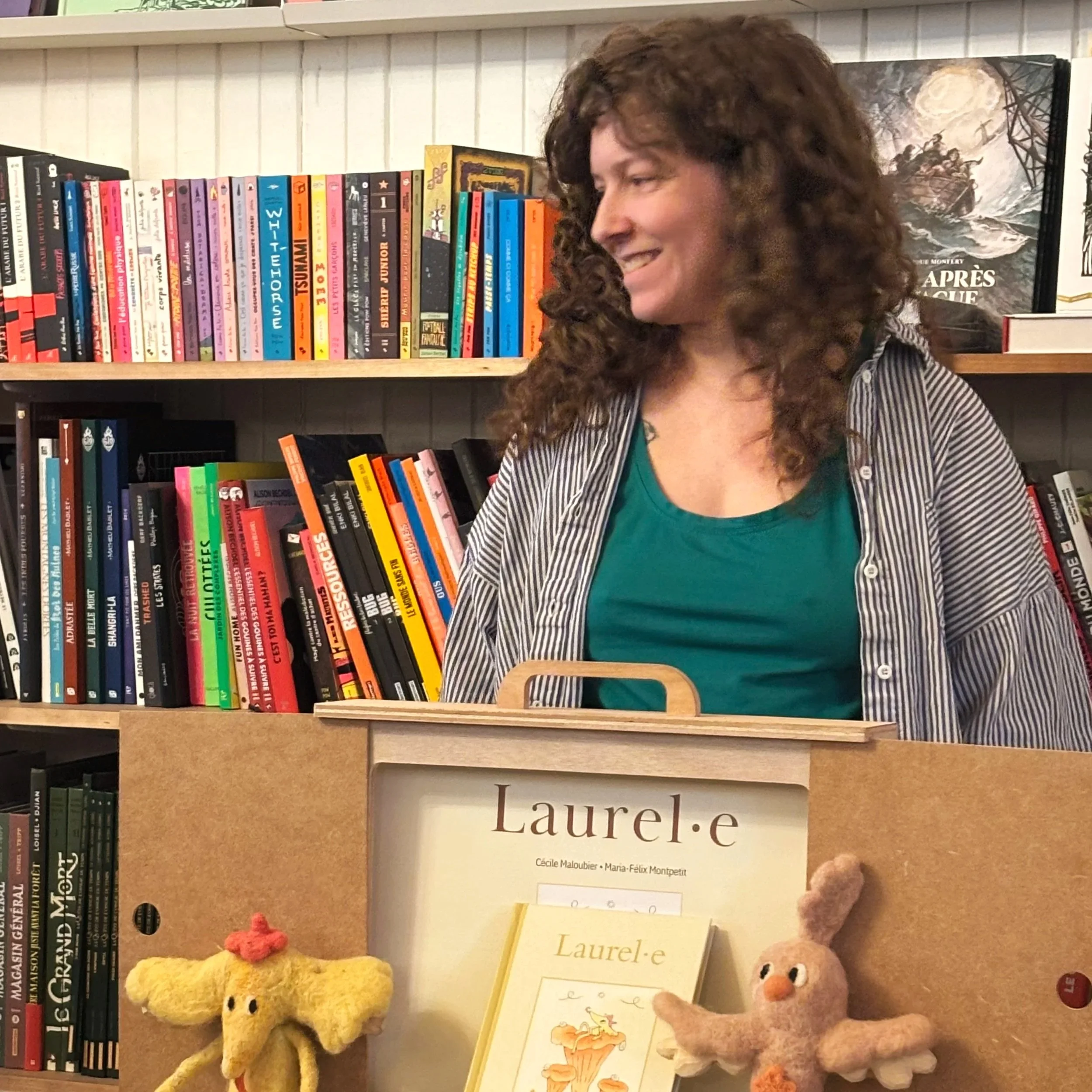 Photo d'une étagère avec des livres colorés et deux peluches, une jaune et une rose, au premier plan. Une femme blonde avec des cheveux bouclés se trouve sur la droite, souriant et regardant vers la gauche. Le livre principal est intitulé 'Laurel.e'.