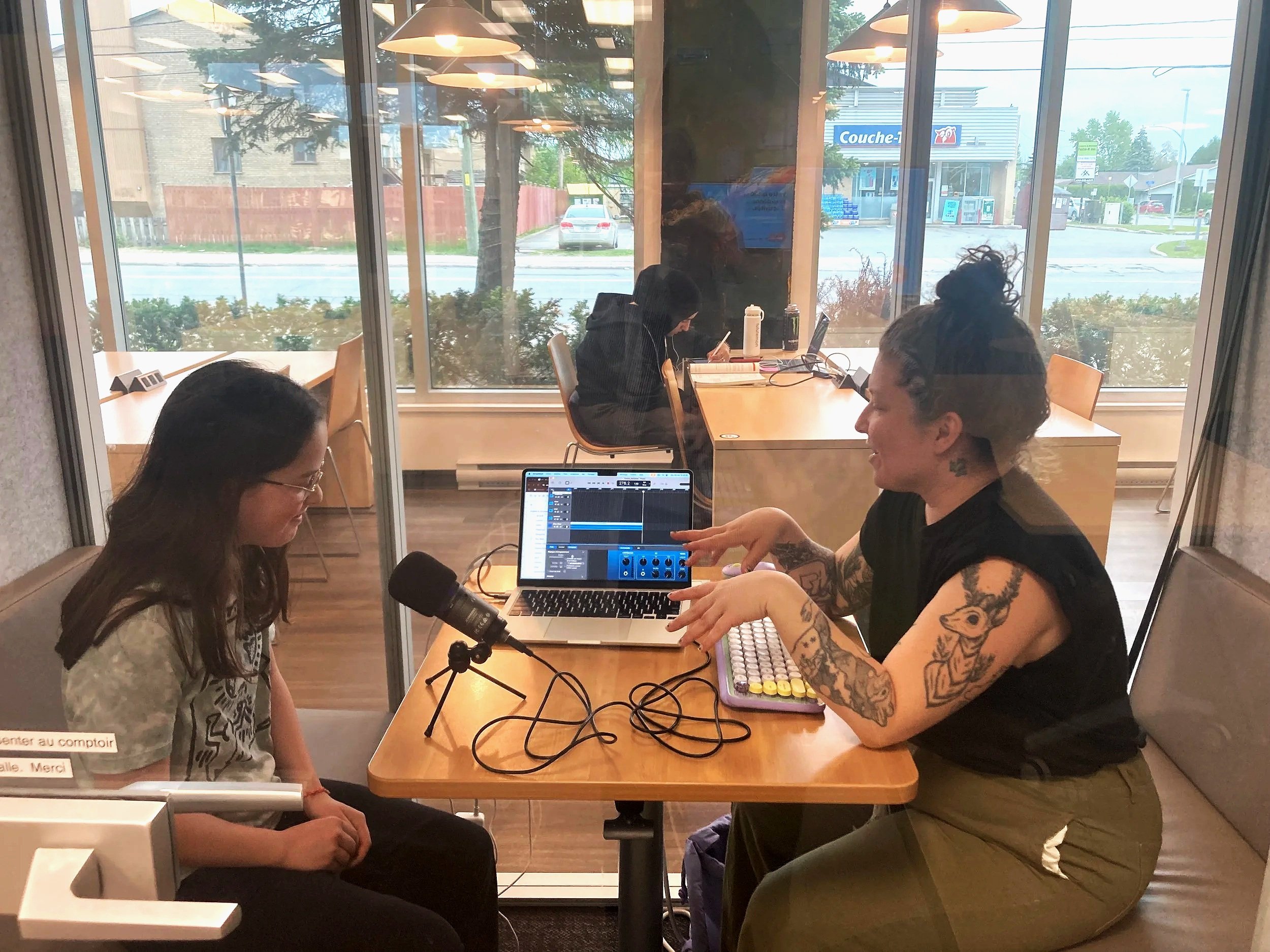 Groupe de personnes dans une salle de réunion, avec deux femmes côtes à côtes, l'une avec des tatouages et l'autre avec des écouteurs, utilisant un ordinateur portable et un microphone, à travers une fenêtre donnant sur une rue avec magasins et voitures.