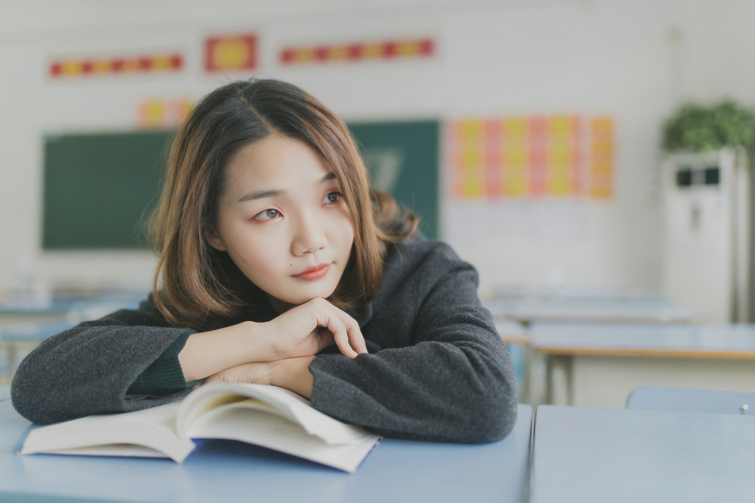 A young woman with shoulder-length brown hair, wearing a dark gray sweater, is sitting at a desk in a classroom, resting her chin on her folded arms, and looking thoughtfully to her right. An open book is on the desk in front of her. The classroom background features a green chalkboard, colorful posters, and a white wall with a potted plant on a shelf.