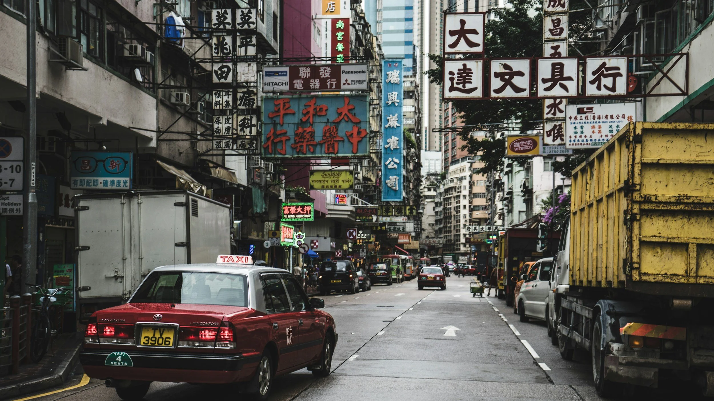 A busy street scene in Hong Kong, with cars, trucks, and taxis, and numerous signs with Chinese characters hanging above the street.