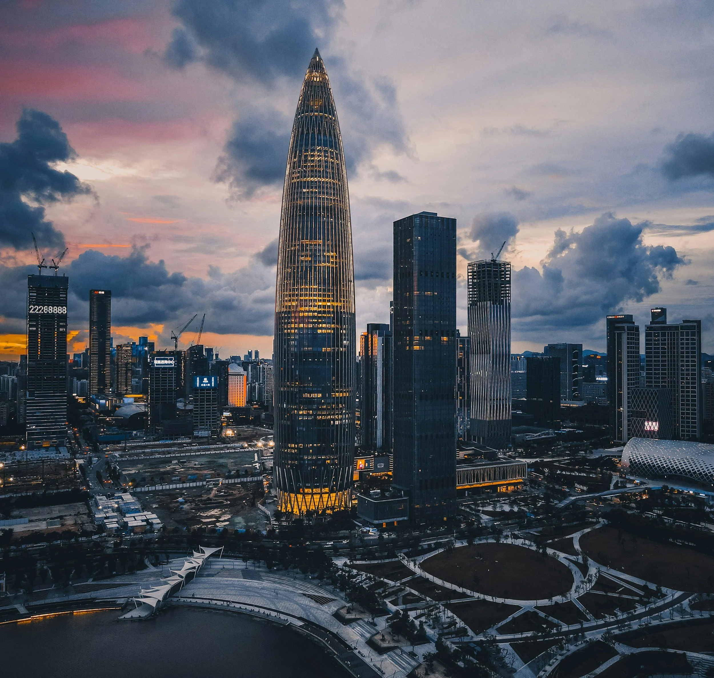 A city skyline at dusk featuring taller buildings, one with a pointed top and illuminated windows, against a cloudy sky with pink and purple hues.
