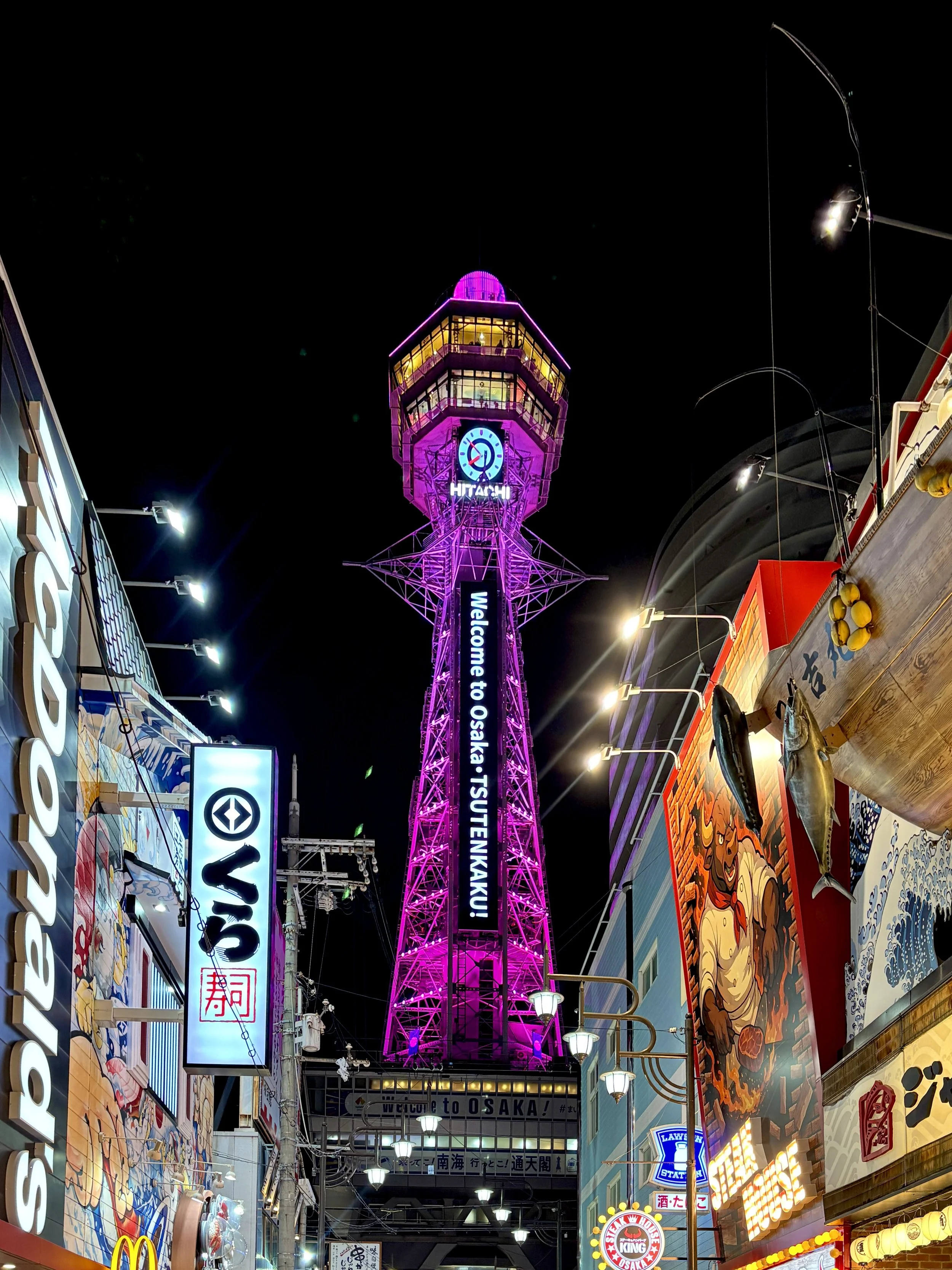 Night view of a colorful urban street in Osaka with neon signs, a lit-up tower with a clock, and various storefronts.