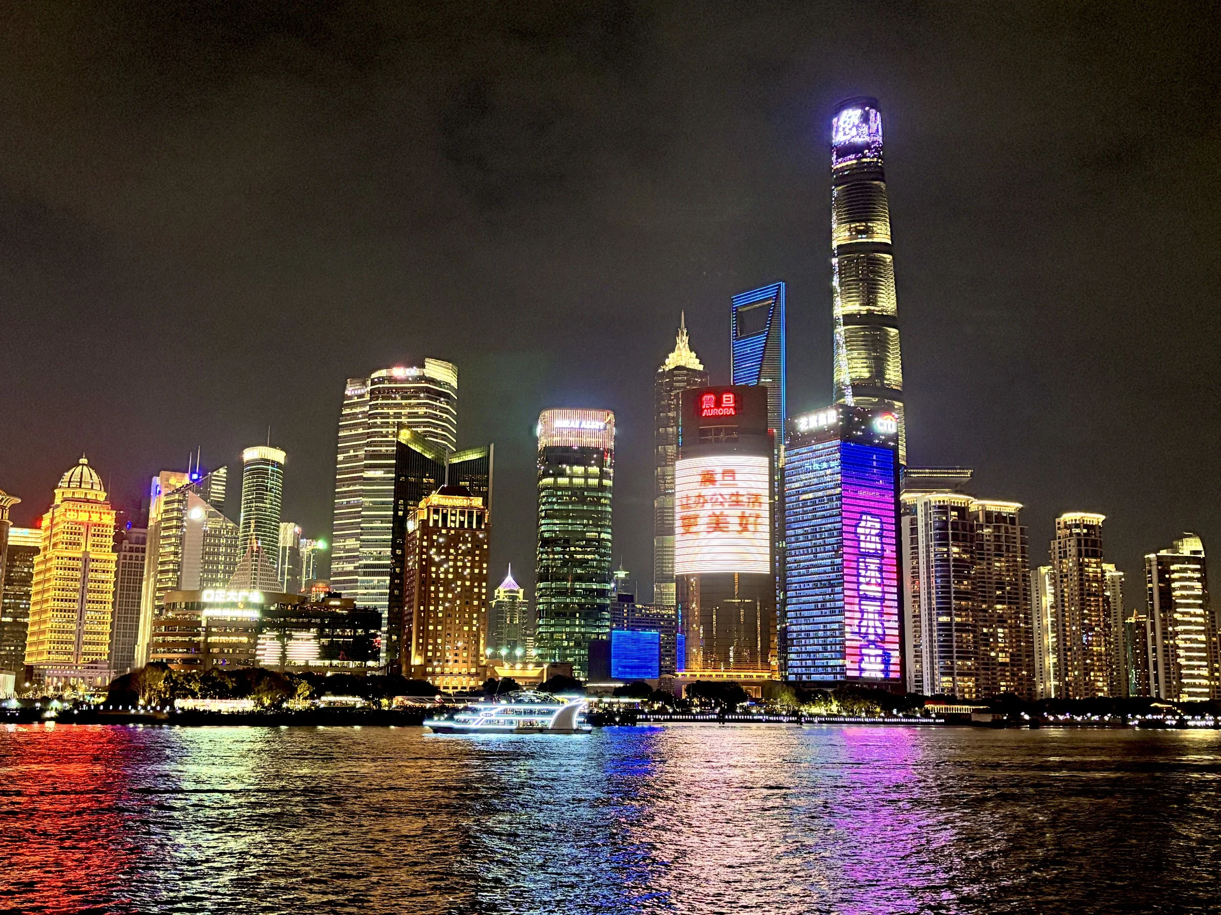 Nighttime city skyline with illuminated skyscrapers along a waterfront, reflected in the water, with a boat passing in the foreground.