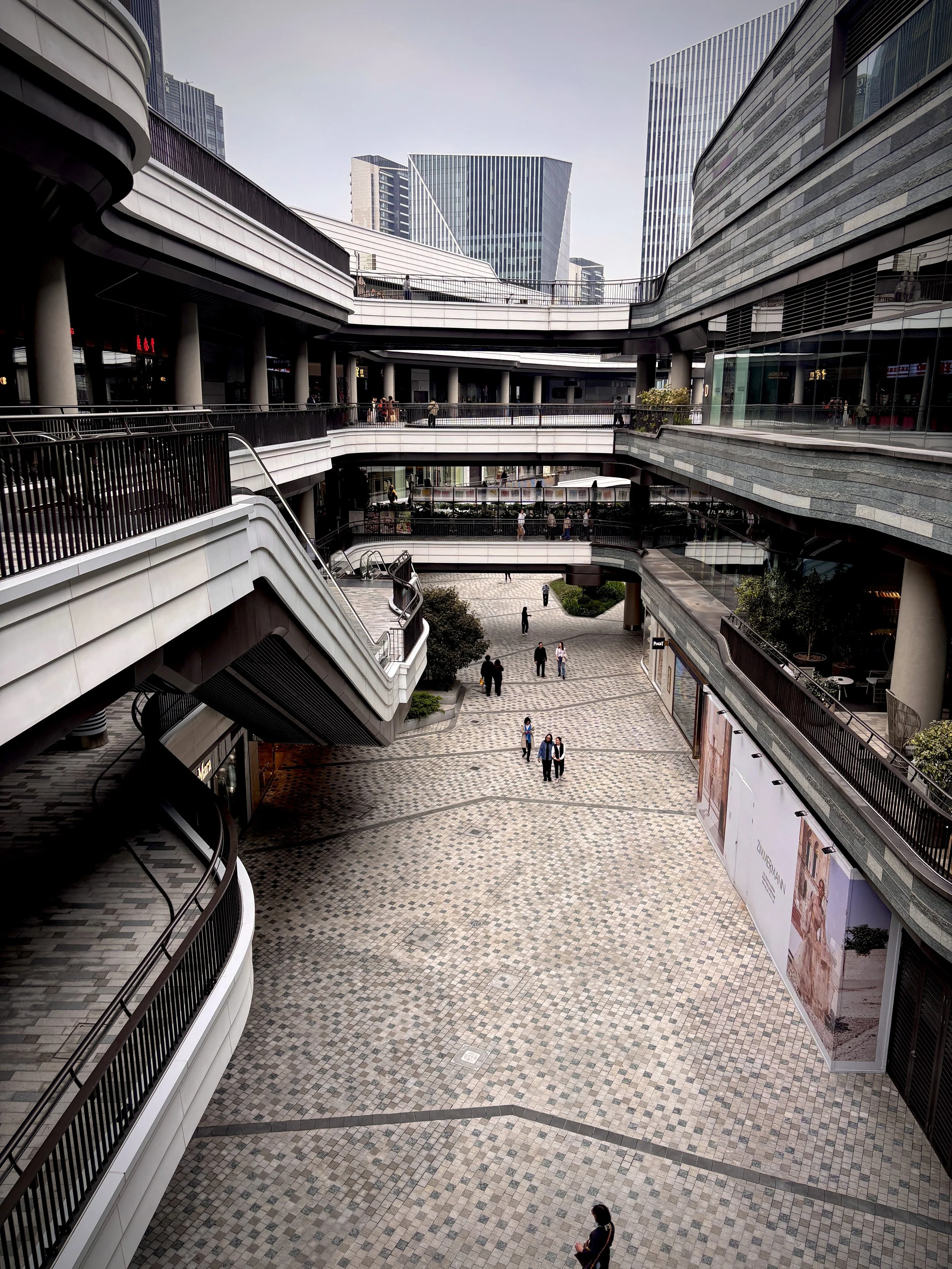Multiple levels of an outdoor shopping mall with curved walkways, glass storefronts, and tall modern buildings in the background. Several people are walking around on the walkways.