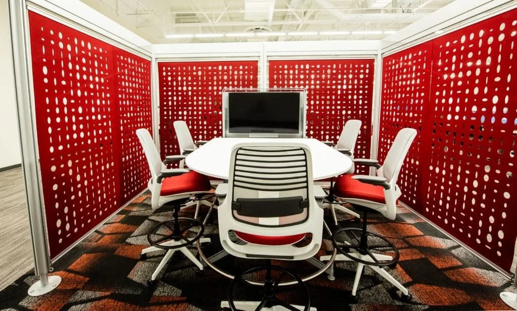 Conference room with red patterned walls, white round table, six white chairs with red cushions, and a large TV monitor.