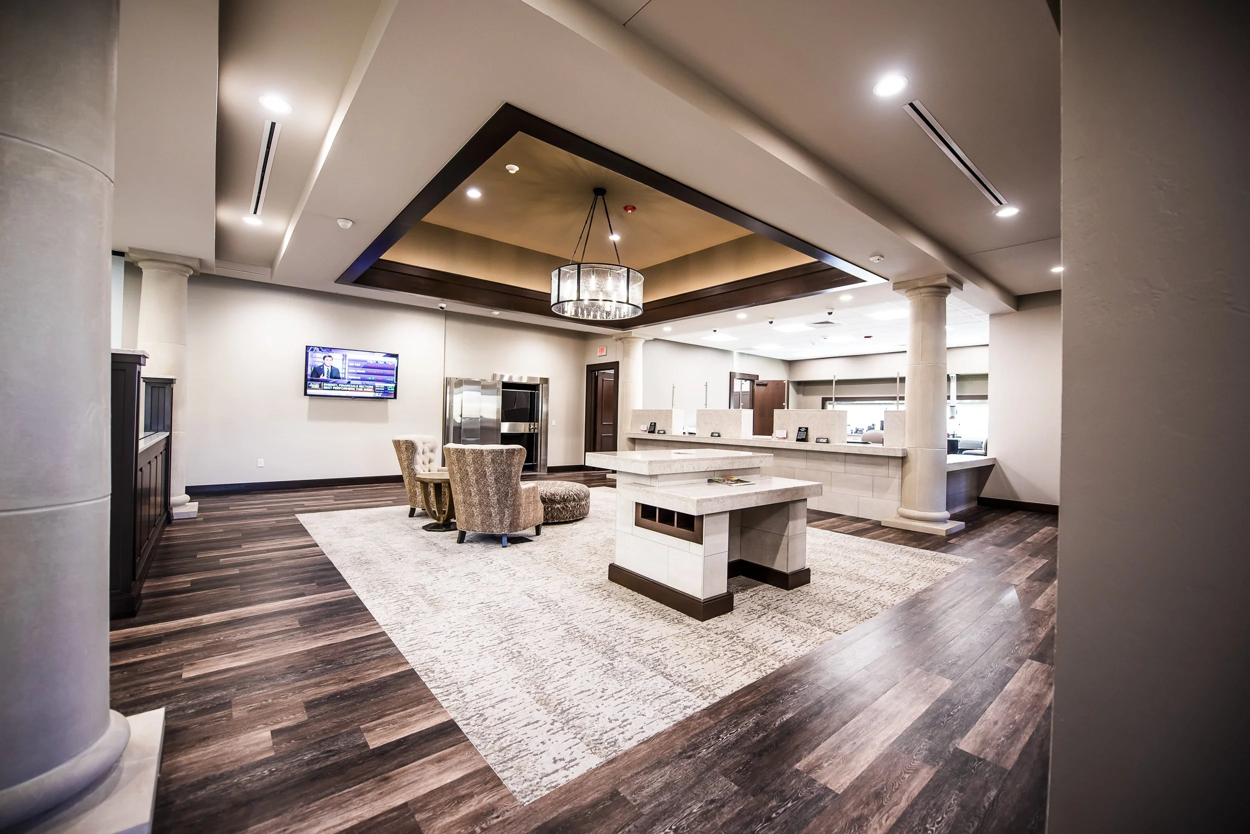 A spacious hotel lobby with dark wood flooring, cream-colored walls, and a large patterned area rug. There are a few beige upholstered chairs around a white marble table, a chandelier hanging from a recessed ceiling, and a television mounted on the wall showing a news channel.