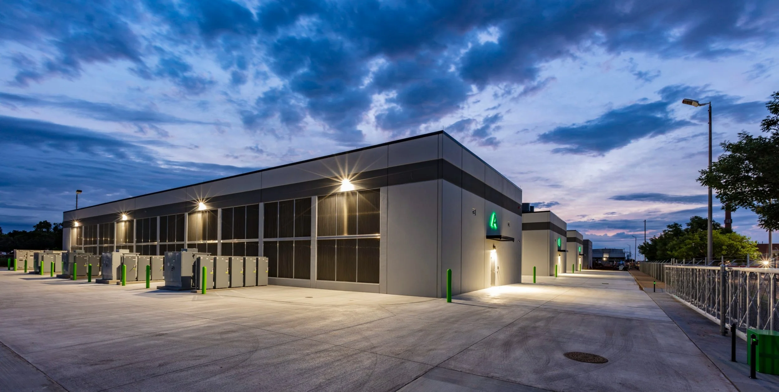 Modern industrial building with solar panels on the roof, illuminated by exterior lights at twilight, surrounded by a paved area with green safety bollards and electric vehicle charging stations.