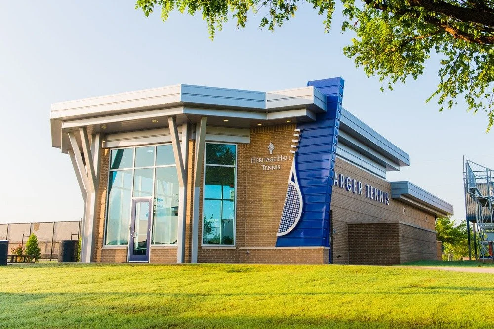 A modern tennis facility with a brick exterior, large glass windows, and a blue facade with a tennis racket graphic. Signage indicates it is the Heritage Hall Tennis and Bigger Tennis. There is greenery and a tree in the foreground, and a bright blue sky.