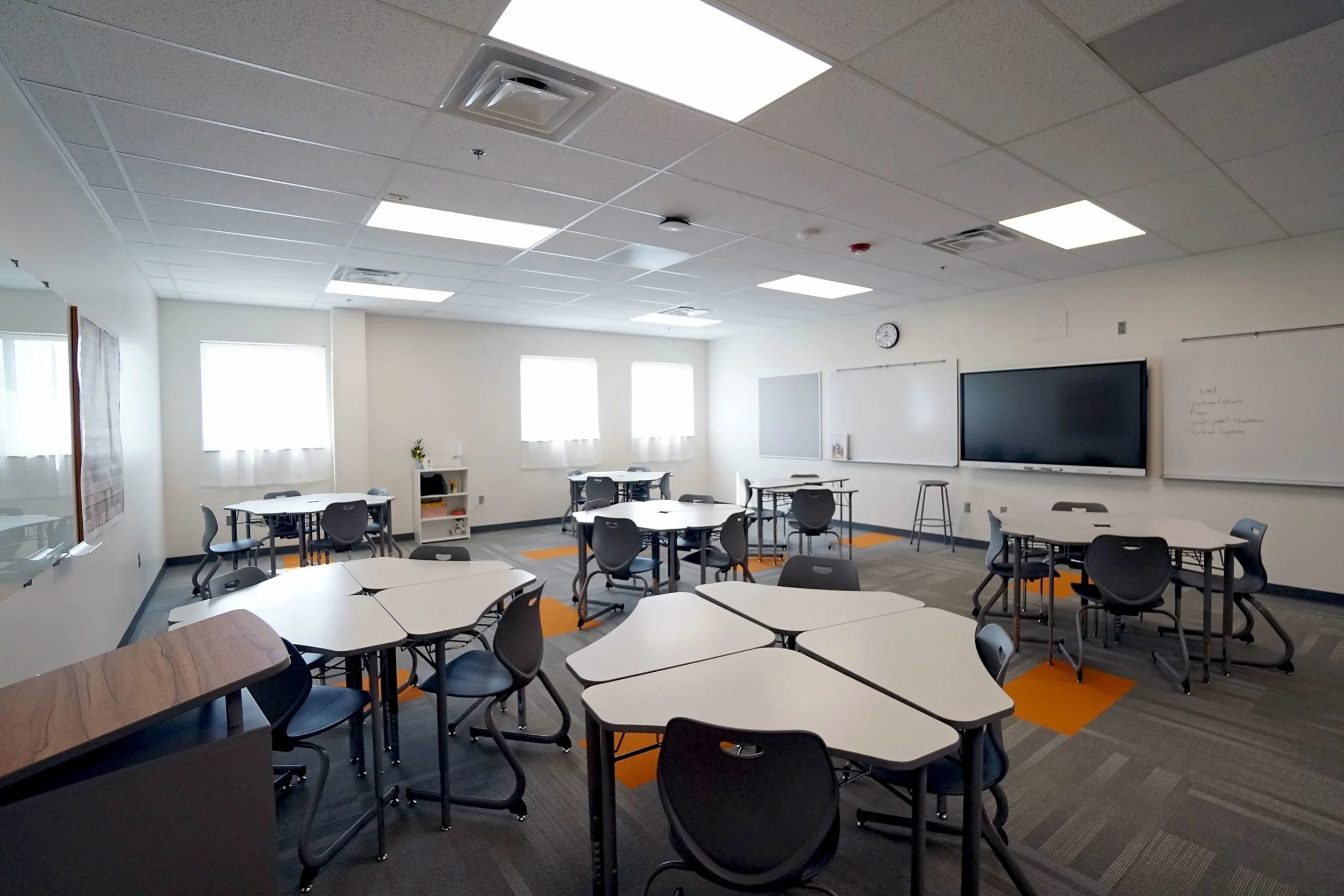 Empty classroom with round tables and chairs, whiteboards, and a large TV screen on the wall. Bright natural light coming through windows, with ceiling lights and a clock above the whiteboards.