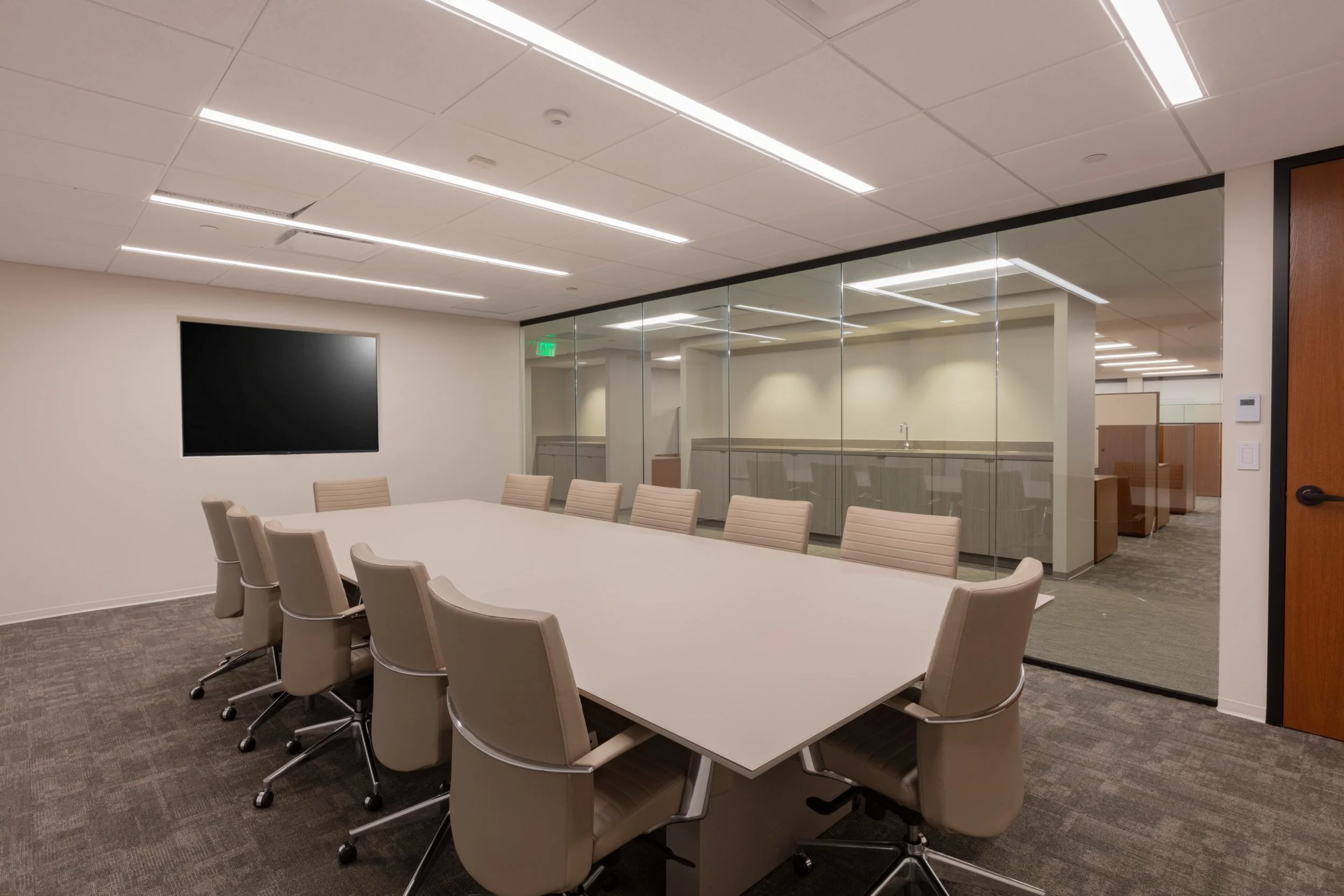A modern conference room with a large oval white table surrounded by beige ergonomic office chairs. The room features a wall with glass partitions, a black wall-mounted display, and a kitchenette with cabinets and a sink in the background. The ceiling has recessed lighting and an access panel.