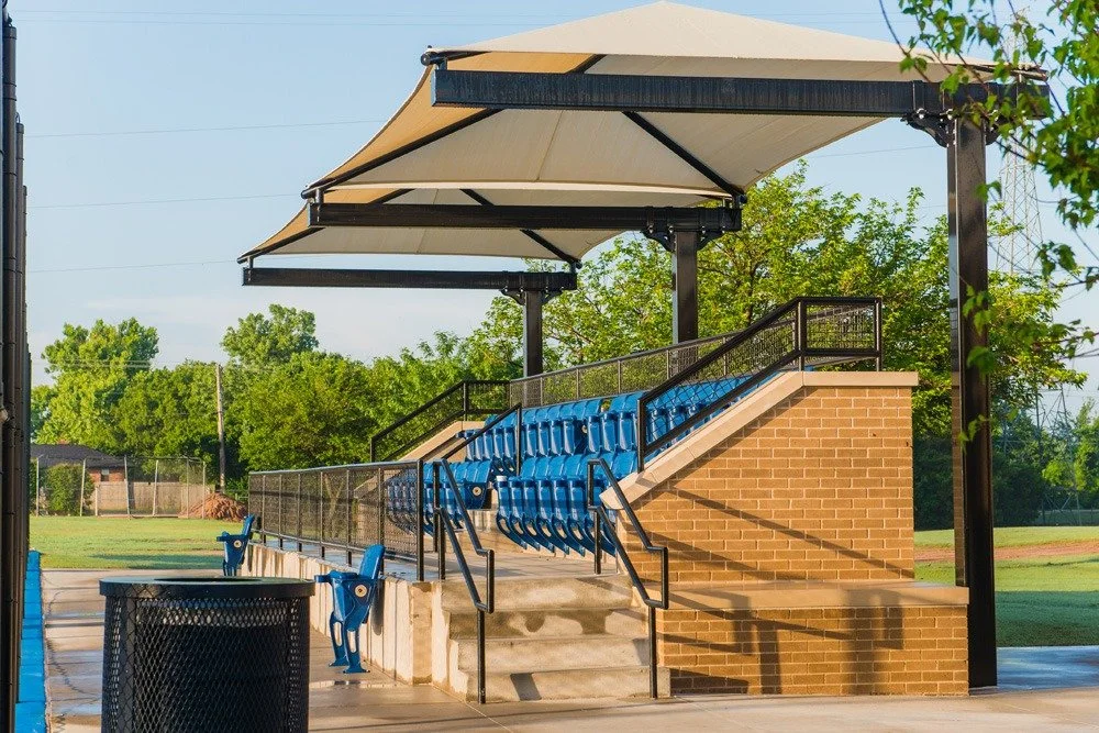 Empty stadium bleachers with blue seats under large shaded canopies and a railing, next to a brick wall and a field with trees in the background.