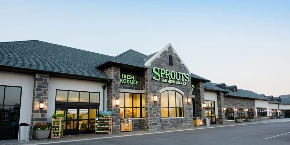 Exterior view of Sprouts Farmers Market grocery store with stone accents, green signage, and outdoor display of produce.