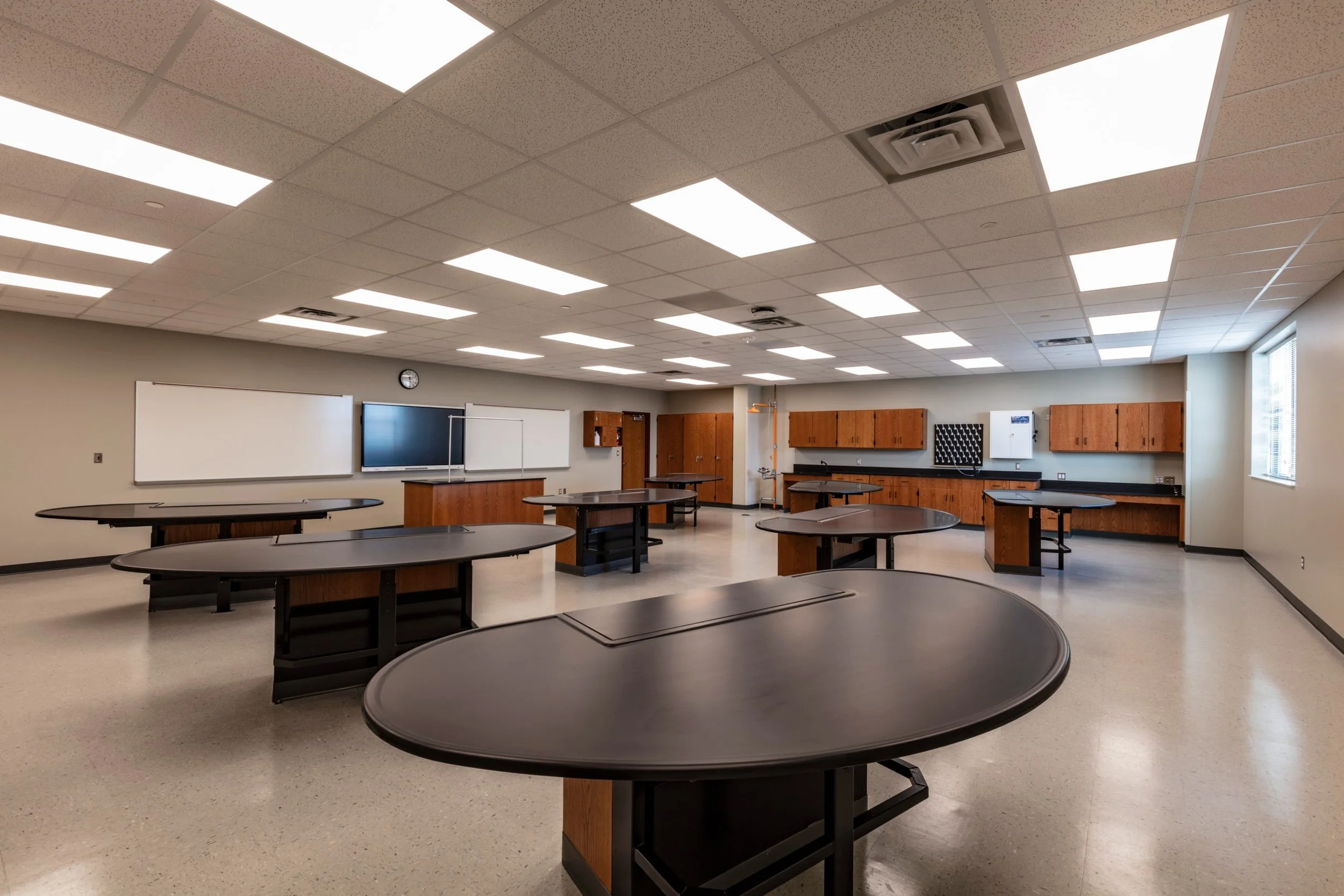 An empty classroom with multiple round desks, wooden cabinets mounted on the wall, whiteboards, and a large window letting in natural light. Ceiling tiles and fluorescent lights are visible.