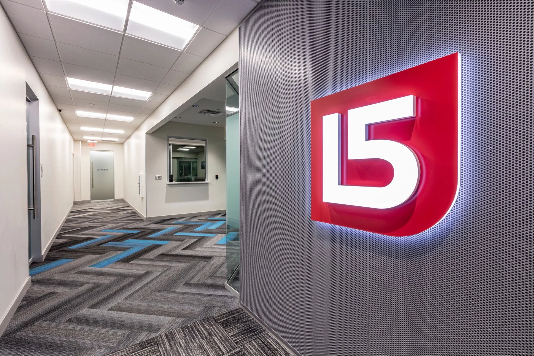 Hallway with modern carpeting, white walls, ceiling lights, a closed door marked 'NOT AN EXIT', and a large illuminated red and white sign with the number 5 on a metallic perforated wall.