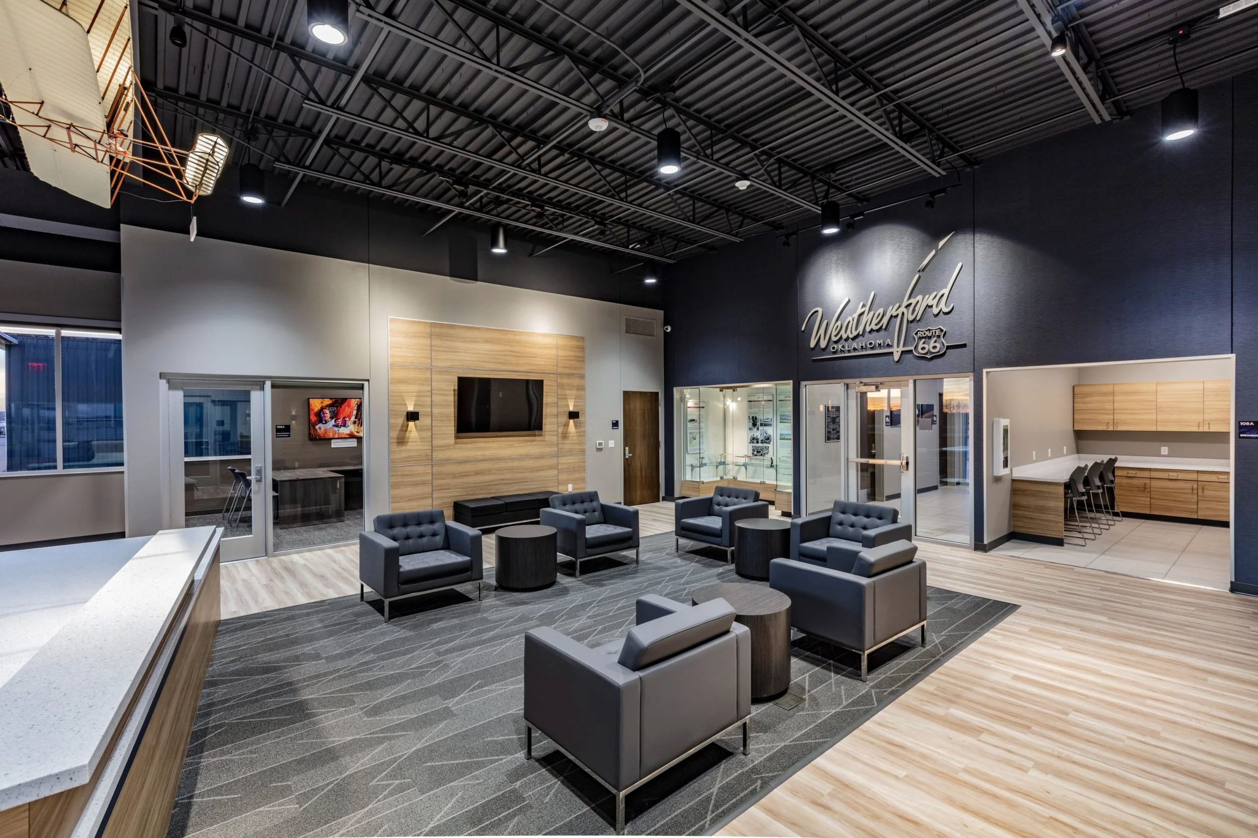 Interior of a modern lobby area with black, gray, and wood accents, featuring several black armchairs, a coffee table, and a wall-mounted TV. Sign on the wall reads 'Weatherford Oklahoma Route 66.'