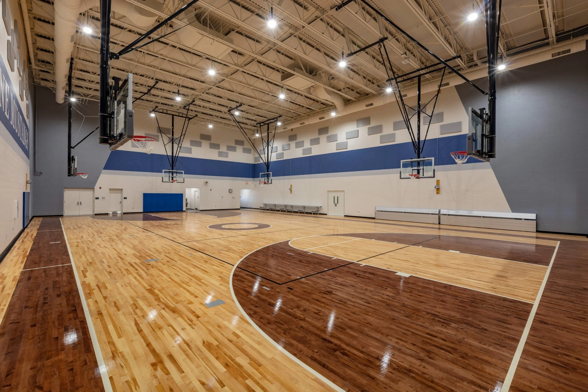 An indoor basketball court with polished wooden flooring, multiple basketball hoops, and overhead lights, in a spacious gymnasium.