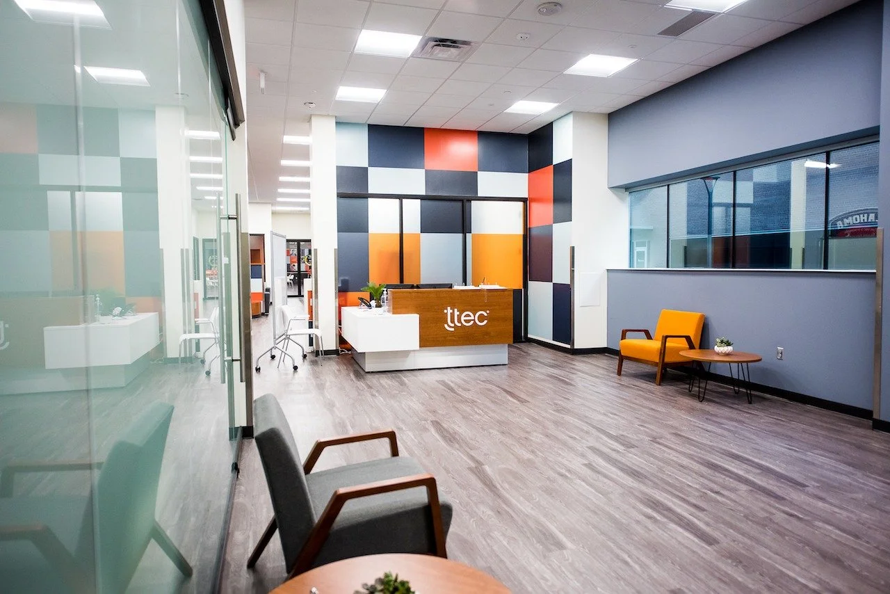 Modern office reception area with a wooden front desk displaying the logo 'tttec', colorful geometric wall art, gray and yellow chairs, a glass wall, and large windows.