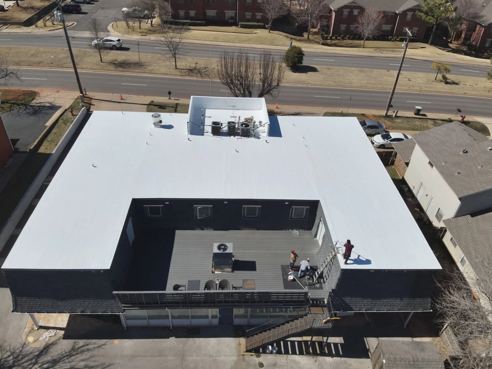 Aerial view of a building's flat roof with workers installing a new white membrane roofing system, with a wooden deck area, and surrounding houses, trees, and a street with parked cars.