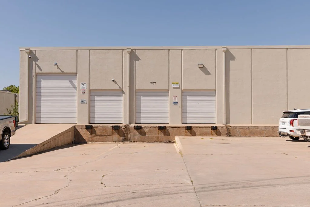 A beige commercial building with three white roll-up garage doors, a small parking lot in front with a few parked vehicles, and a clear blue sky overhead.