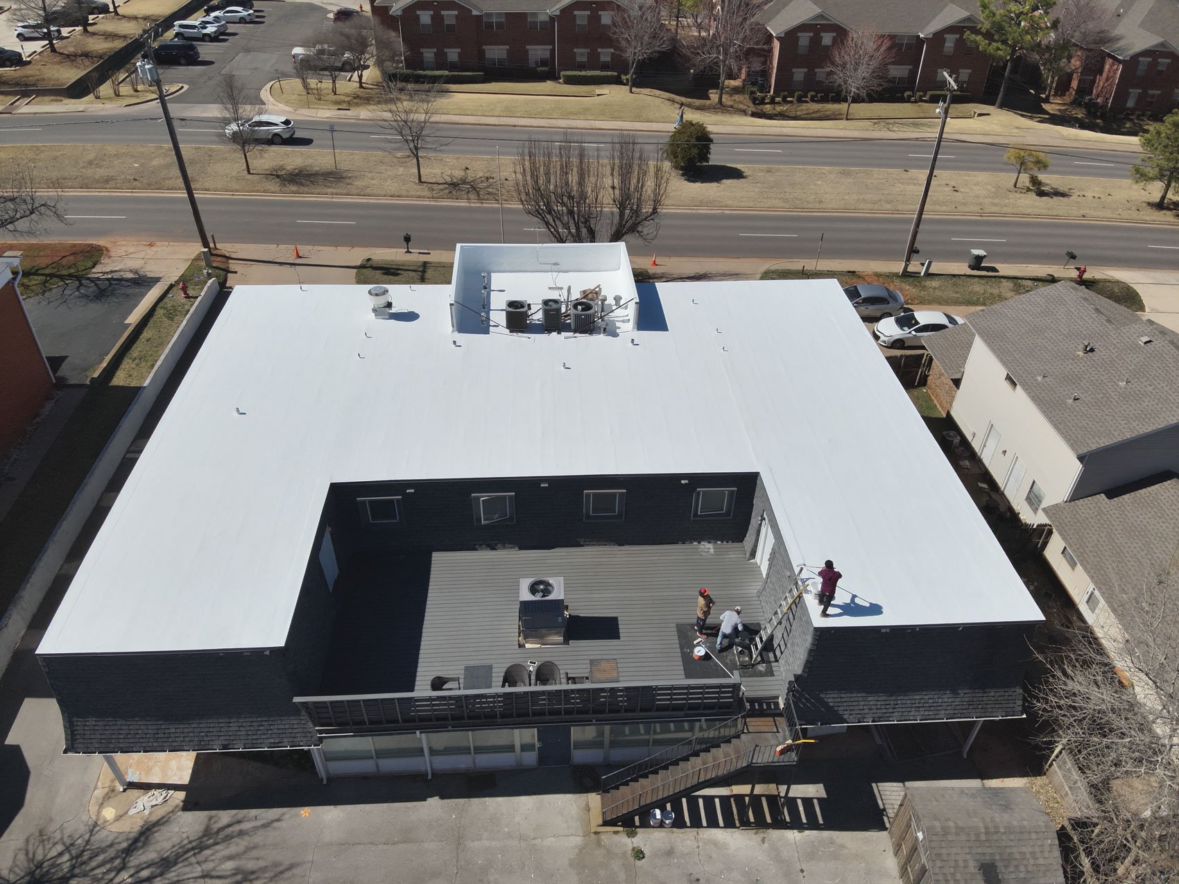 Aerial view of a rooftop under construction or renovation with workers, nearby streets, parked cars, and surrounding residential buildings.