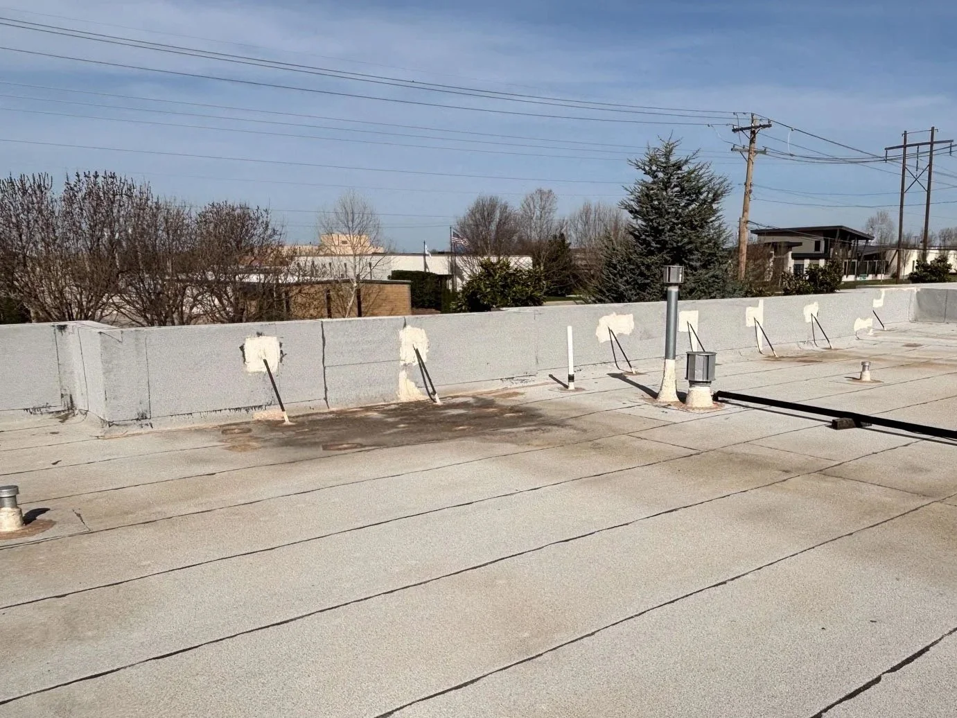 View of a flat rooftop with a low concrete parapet wall, several roof vents, and a small light fixture. In the background, trees, power lines, and buildings are visible under a partly cloudy sky.