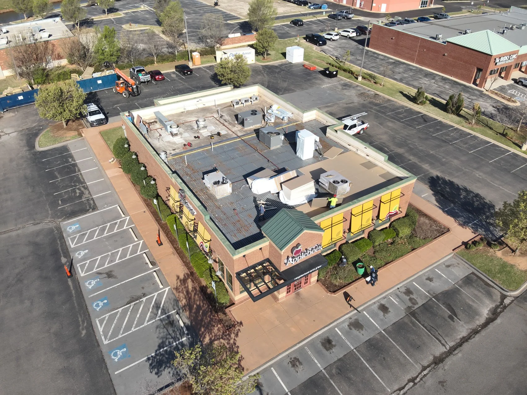 An aerial view of a commercial building with a roof under construction, workers installing roofing materials, and surrounding parking lot with empty parking spaces.