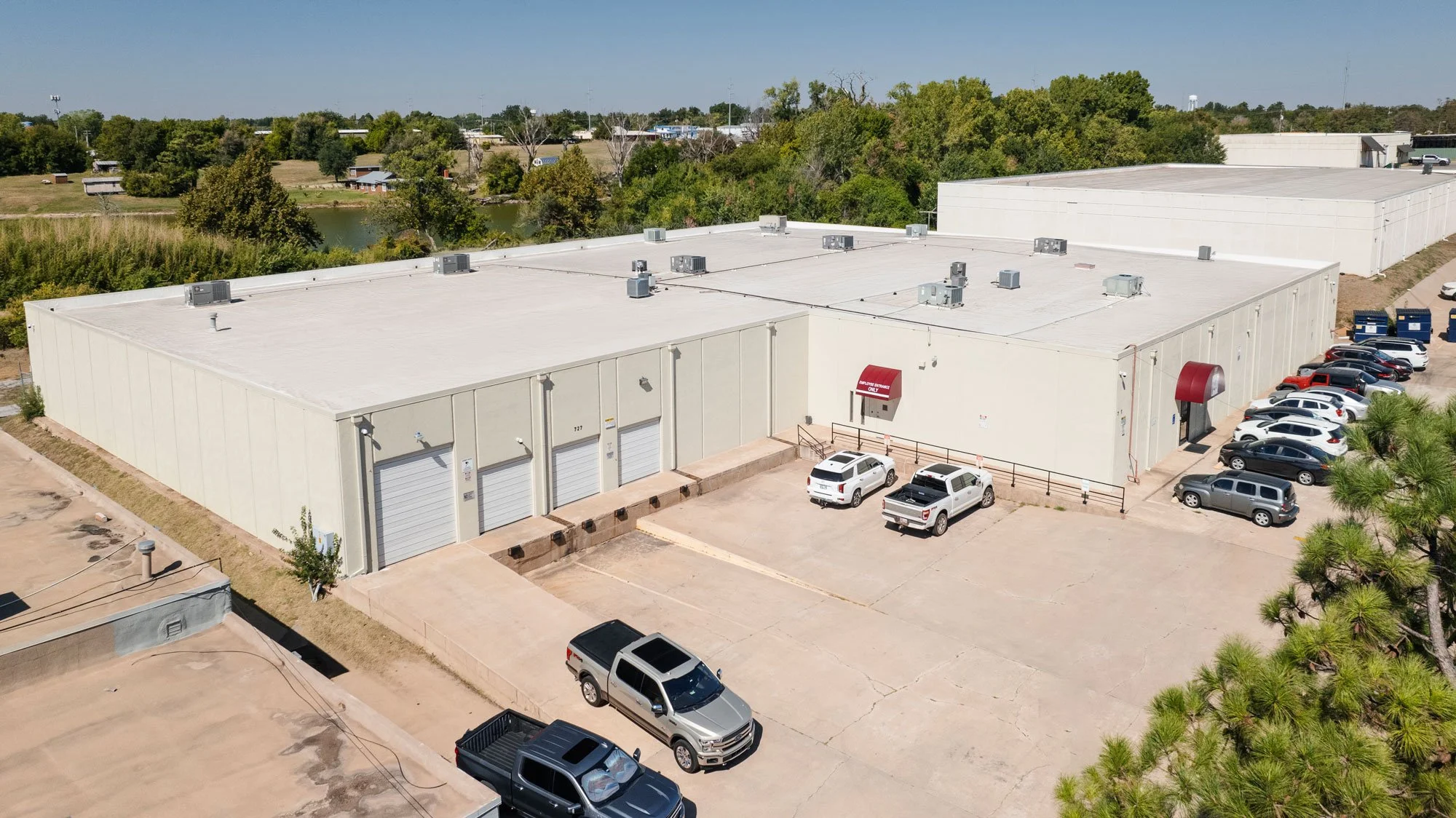 A large commercial or industrial warehouse building with multiple garage doors and parked vehicles in front, situated in a semi-rural area with trees and a water body in the background.