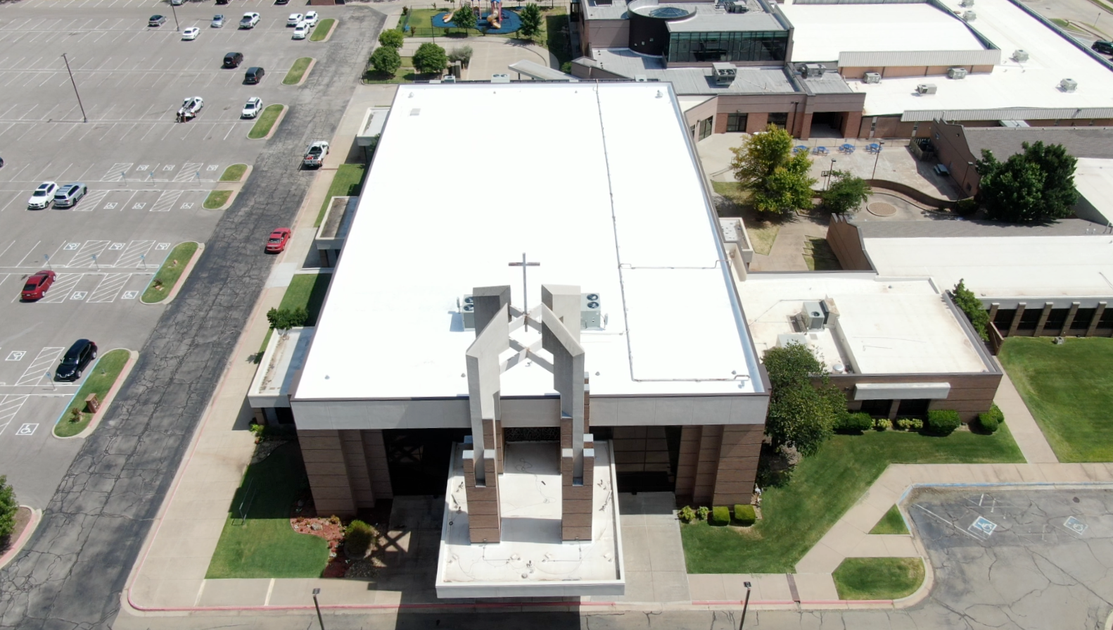 Bird's eye view of a church with a cross on its roof, surrounded by parking lot and adjacent buildings, with trees and green spaces.