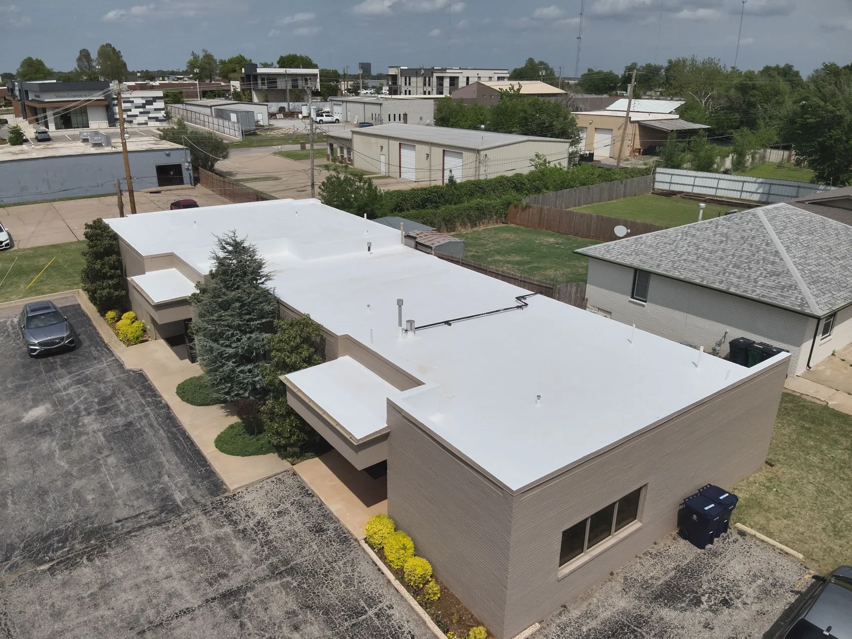 Aerial view of a commercial building with a white flat roof, surrounded by parking lot, trees, and neighboring buildings.