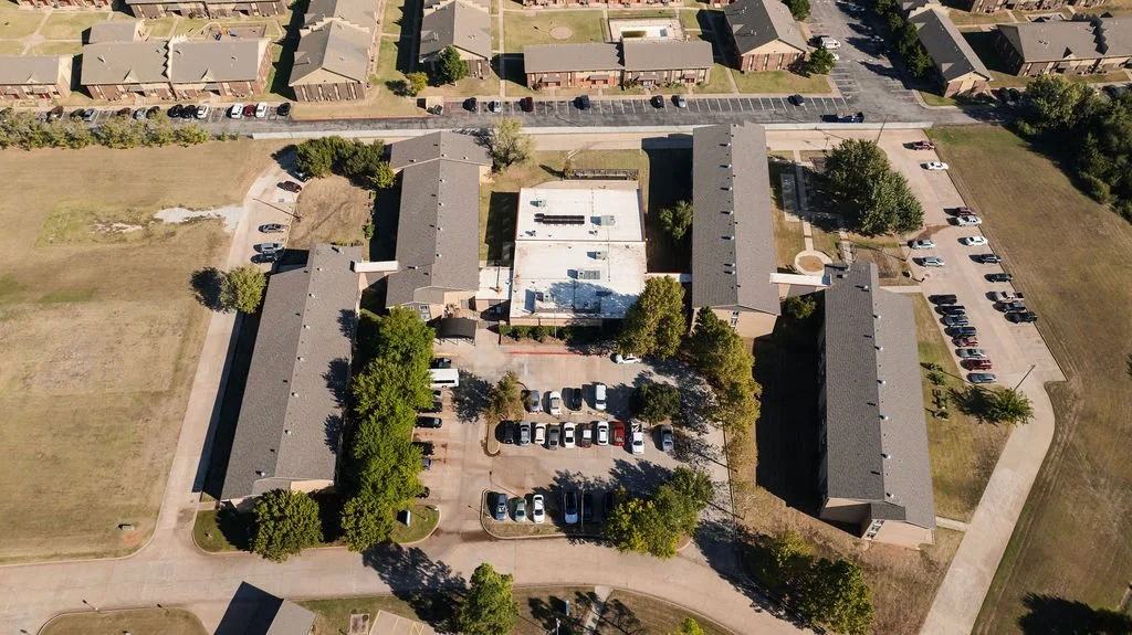 Aerial view of a U-shaped apartment complex surrounded by parking lots, grassy areas, and trees.