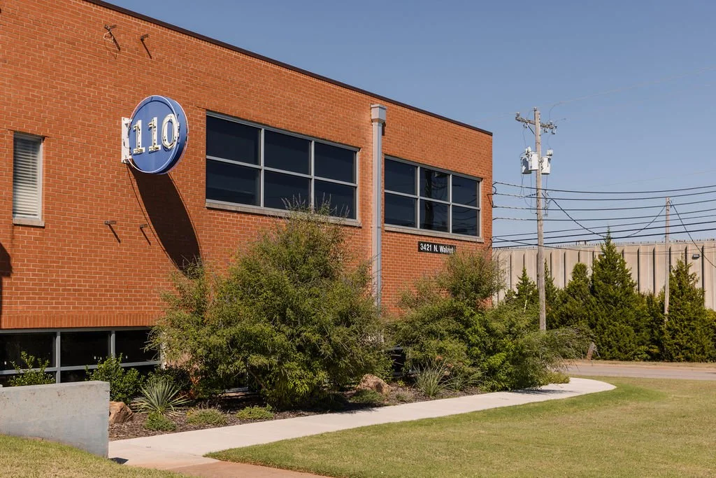 A modern brick building with a circular blue sign displaying the number 110, and a street address sign that reads 3421 N. Wabash, with bushes, trees, and a concrete sidewalk in front.