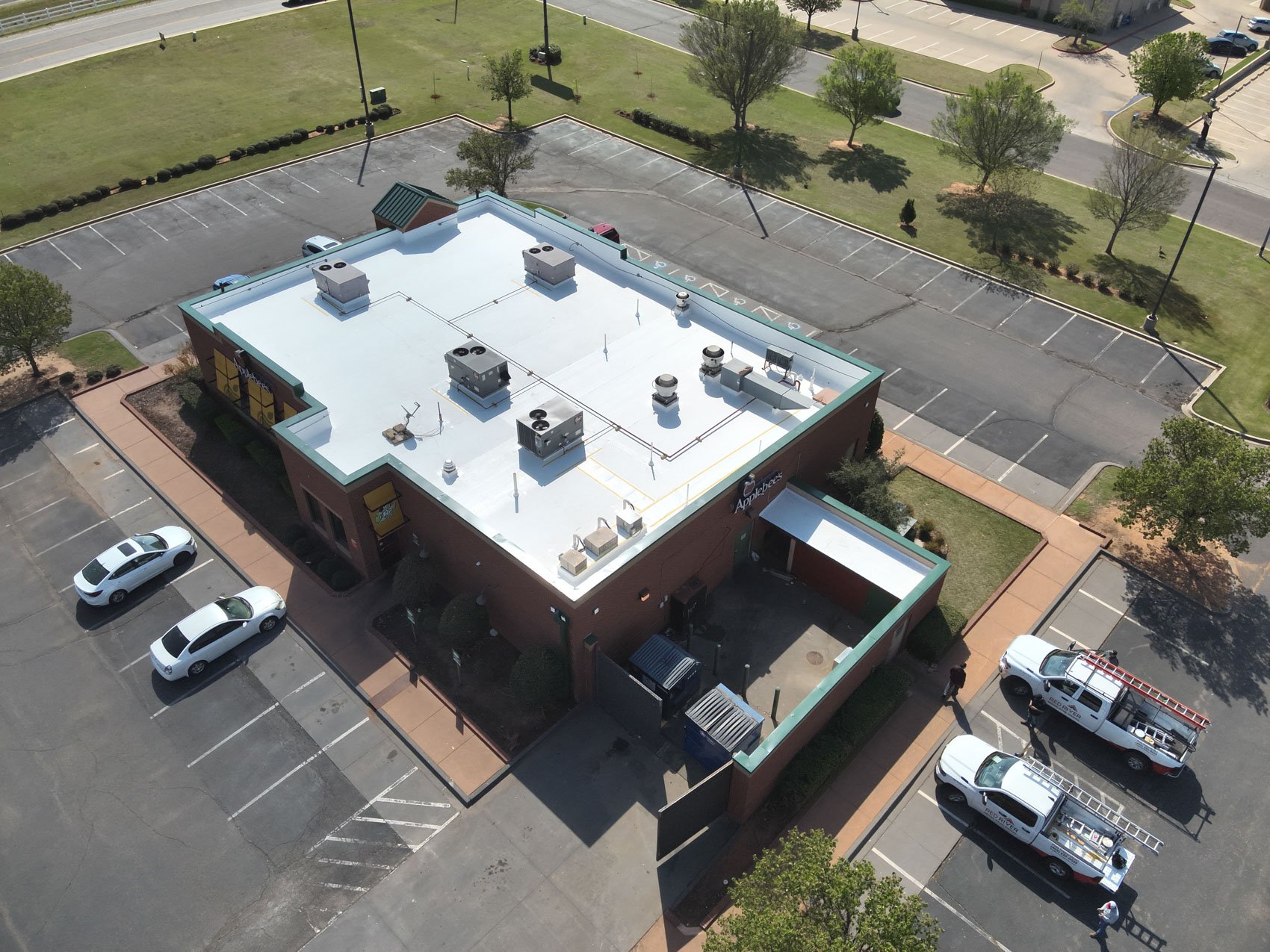 An aerial view of a shopping center with a large parking lot, some parked cars, and a single-story retail store with a white flat roof surrounded by trees and greenery.