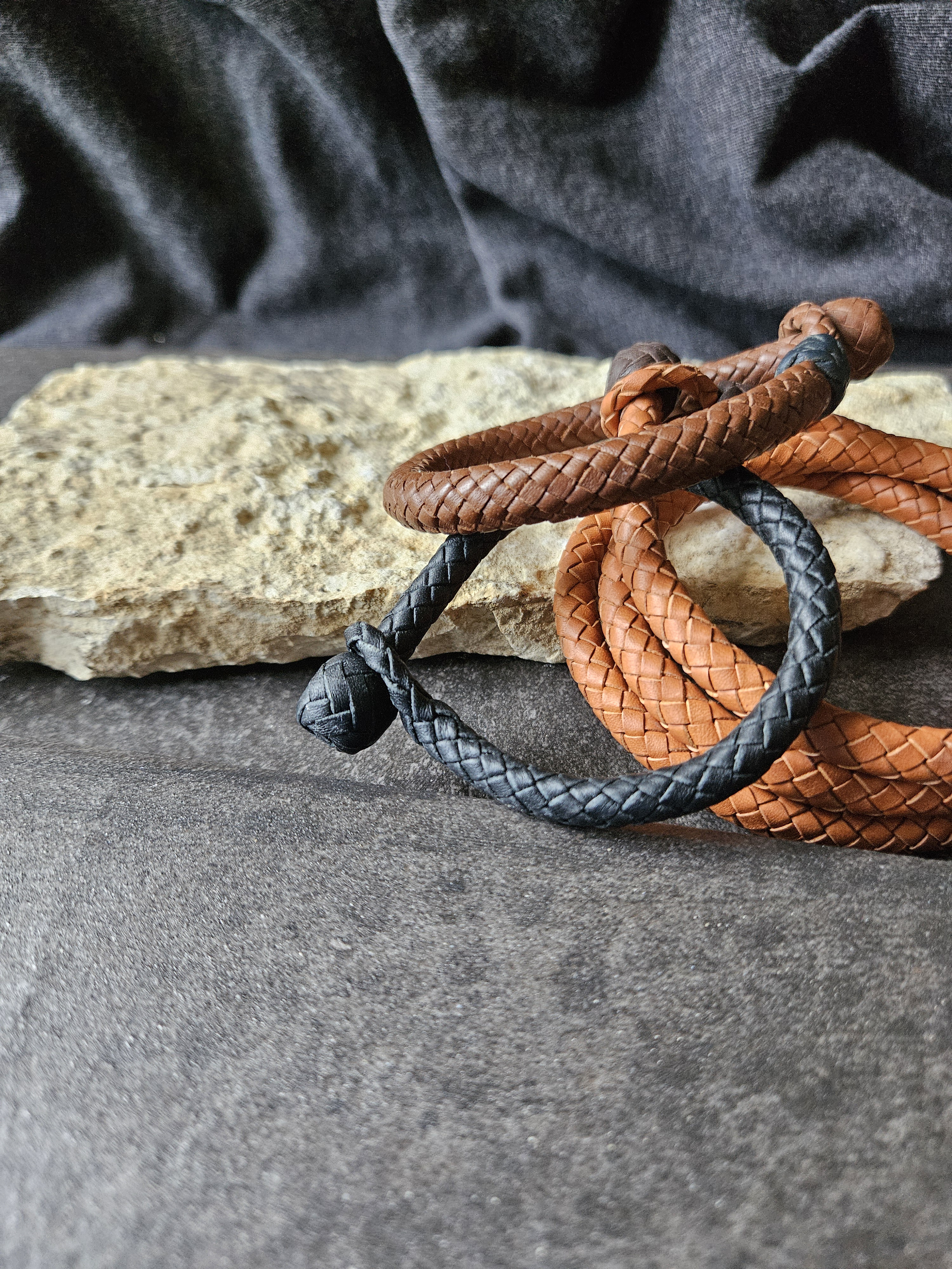 Three braided leather bracelets in black, brown, and tan colors, intertwined and resting on a rough stone surface with a black fabric backdrop.