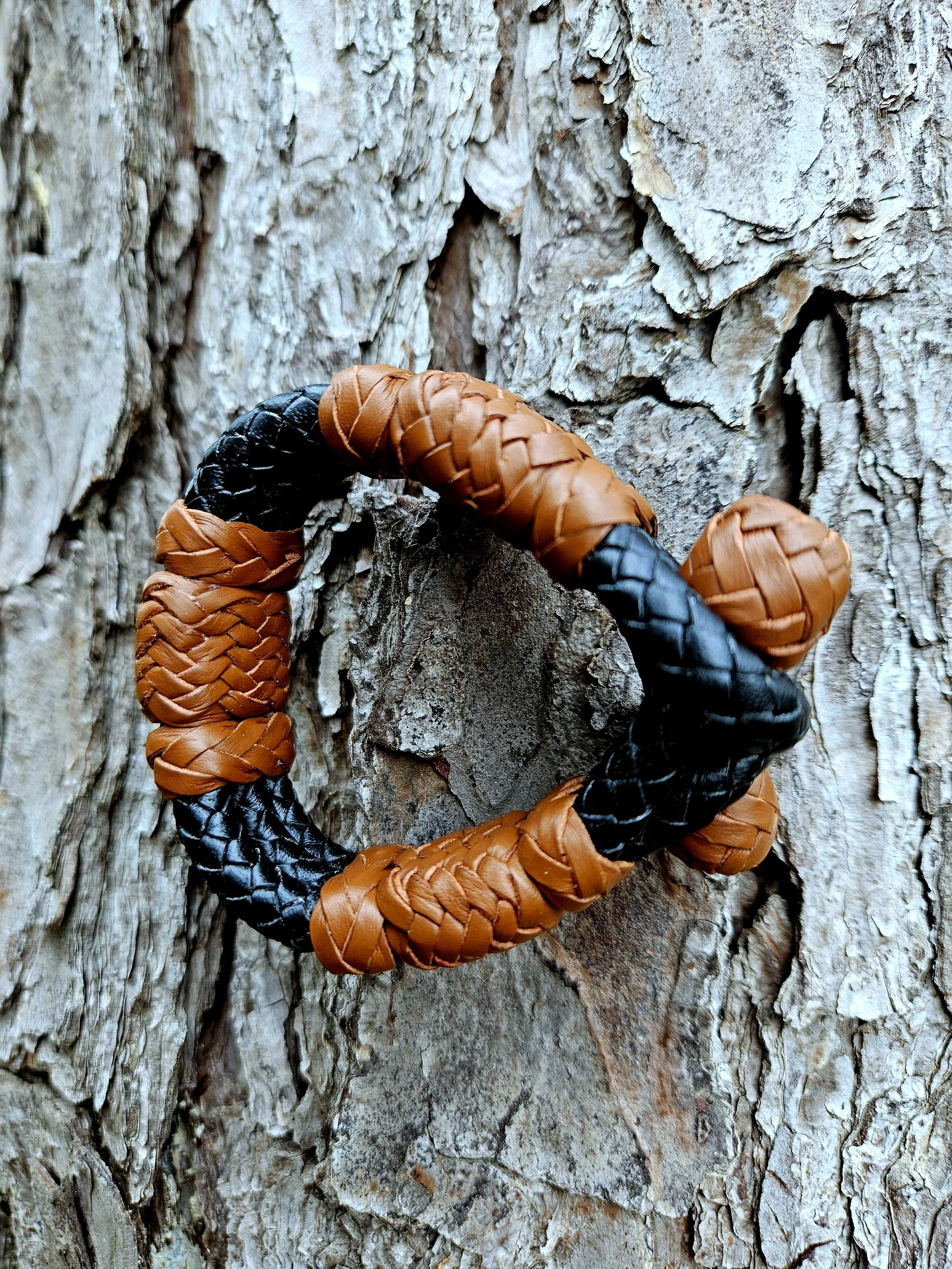 Close-up of a bracelet with orange and black woven straps hanging on the textured bark of a tree.