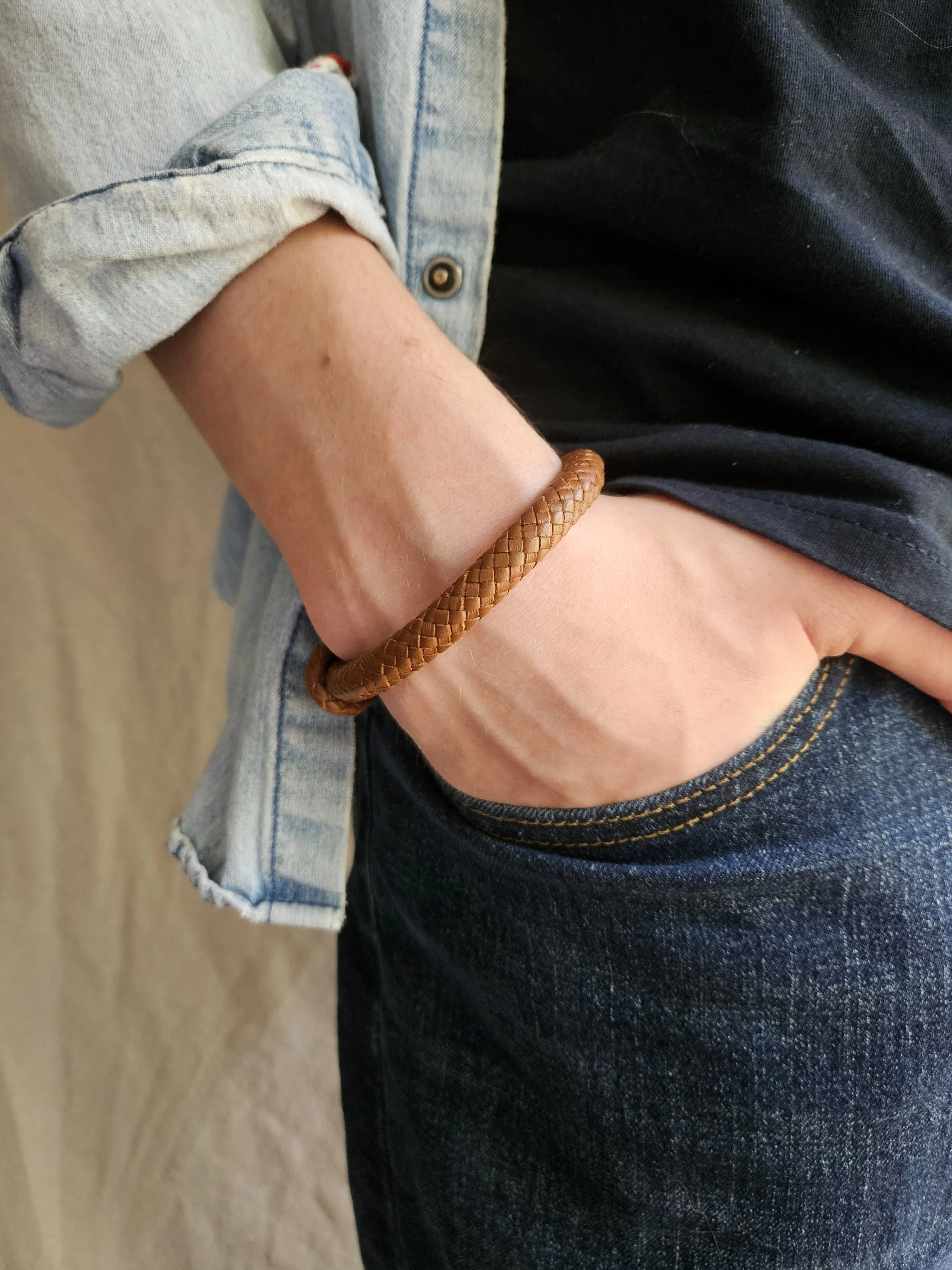 Close-up of someone's arm with a brown leather bracelet, wearing a light-wash denim shirt with rolled-up sleeve, blue T-shirt, and dark blue jeans.