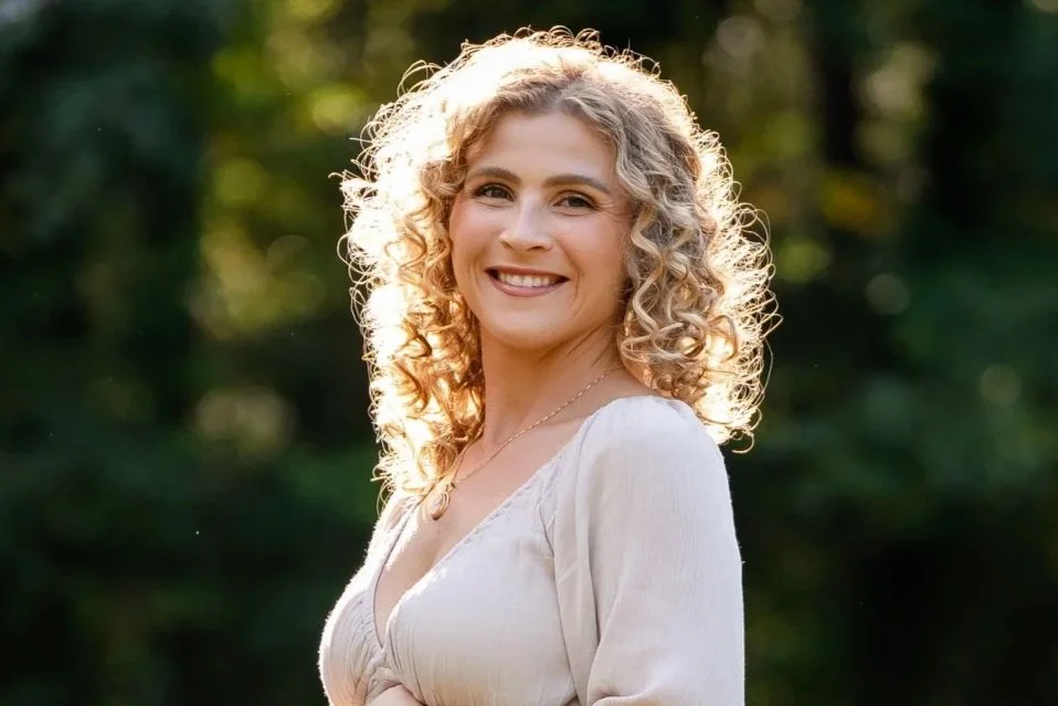 A woman with curly blonde hair smiling outdoors with greenery and sunlight in the background.