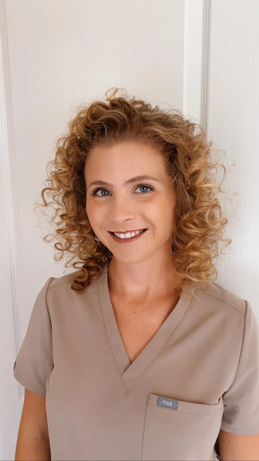 A smiling woman with curly red hair, wearing a beige scrubs top, standing in front of a white door.