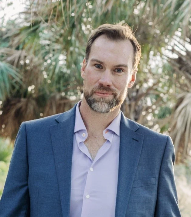 A man in a blue suit jacket and light purple dress shirt standing outdoors with palm trees in the background.
